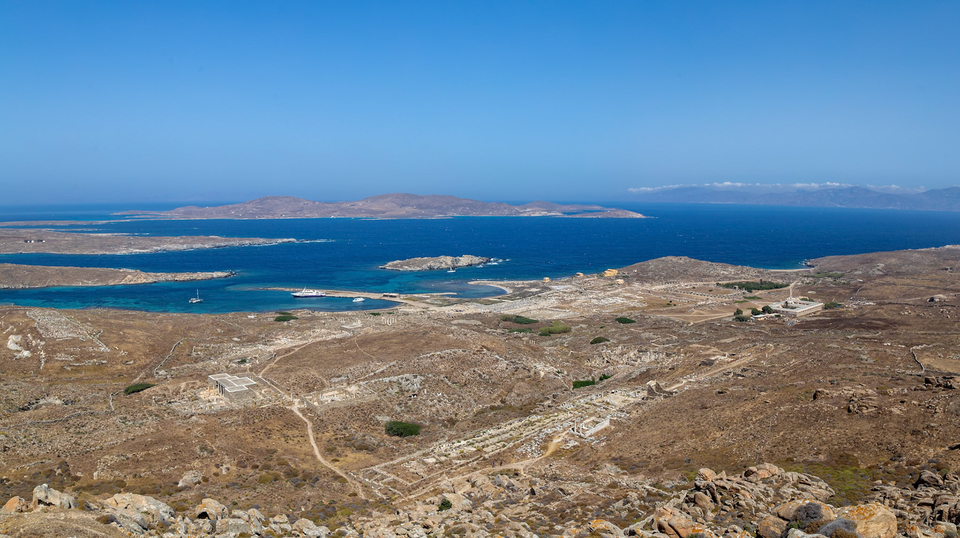 Delos, Greece - May 22nd 2018: A high-angle view shows the ancient ruins of Delos, a Greek island, with the Aegean Sea in the background. Tourists explore the historical site.
