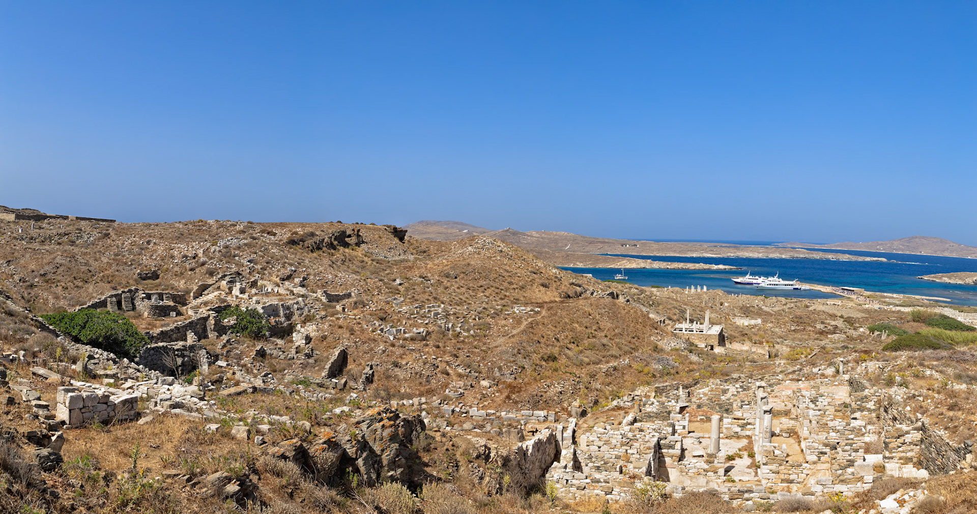Delos, Greece - May 22nd 2018: Ruins of ancient structures dot the landscape, with ferries bringing tourists to explore the historical site.
