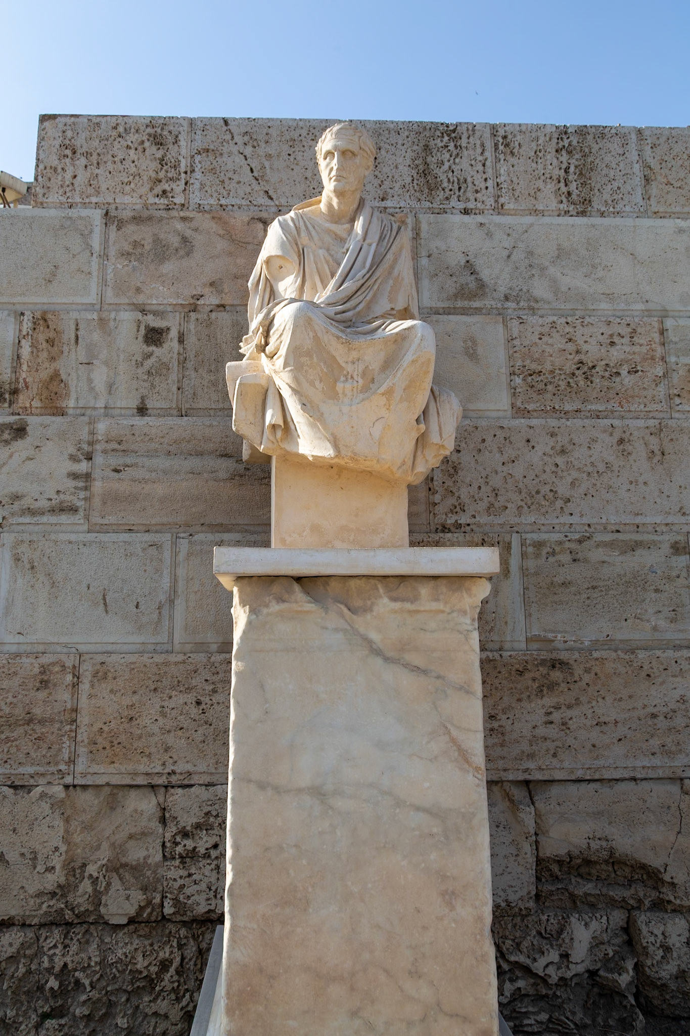 Acropolis, Athens, Greece - May 23rd 2018: A statue of a seated man in a toga sits atop a stone pillar, likely a depiction of a prominent figure.