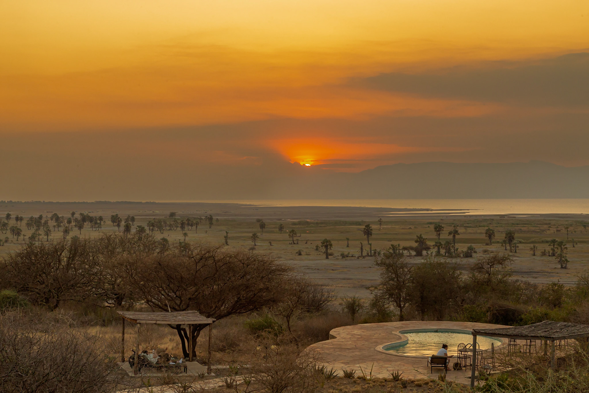 Lake Eyasi, Tanzania - September 27th 2025: A lone figure relaxes by a pool at sunset, overlooking a vast savanna landscape.