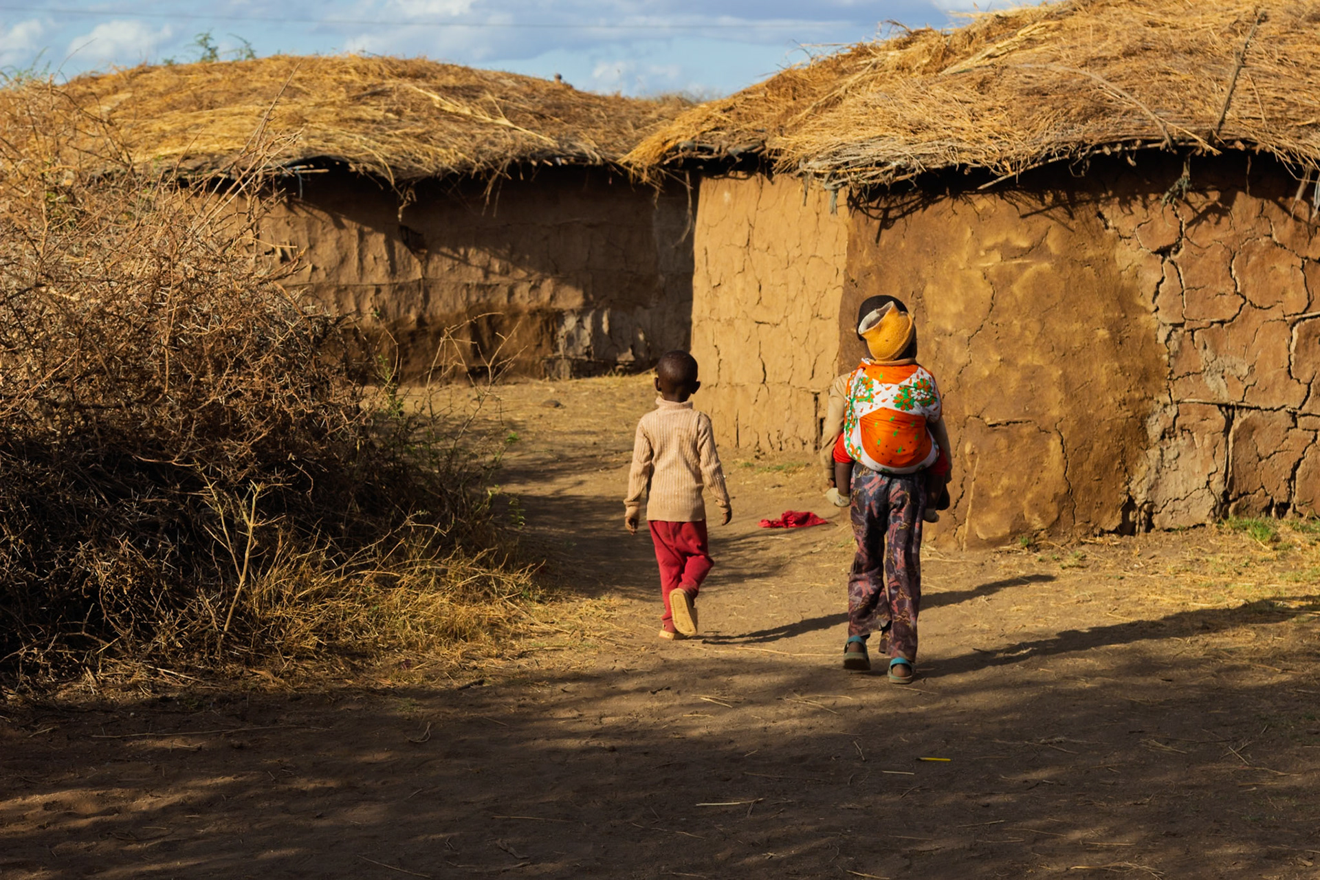 A Maasai mother carries her child on her back as she and another child walk through their village in Kenya.