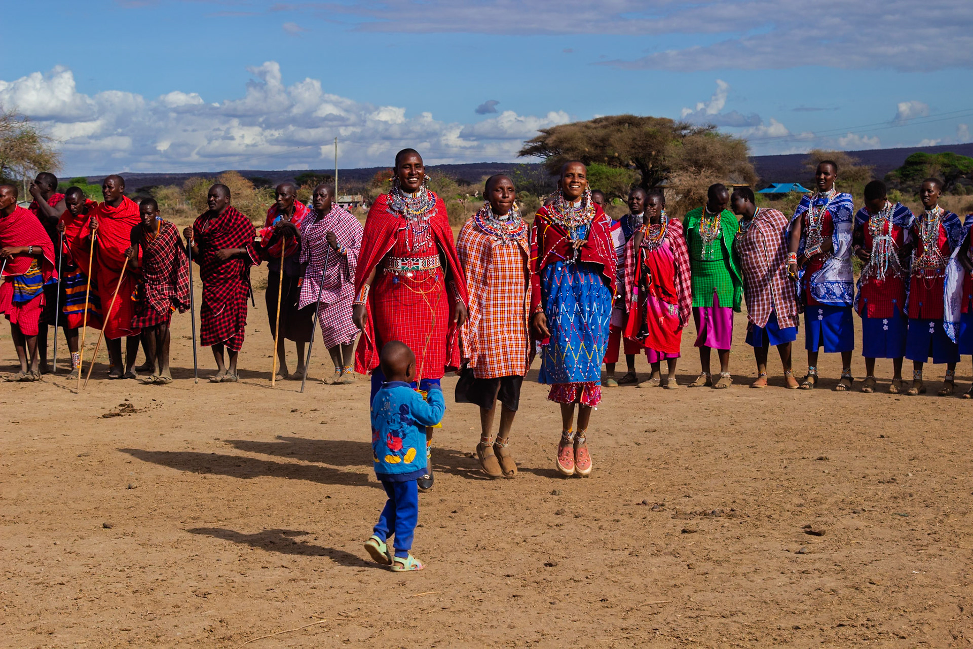 Maasai people in Kenya are seen in traditional dress, with women jumping in unison, while a child looks on.
