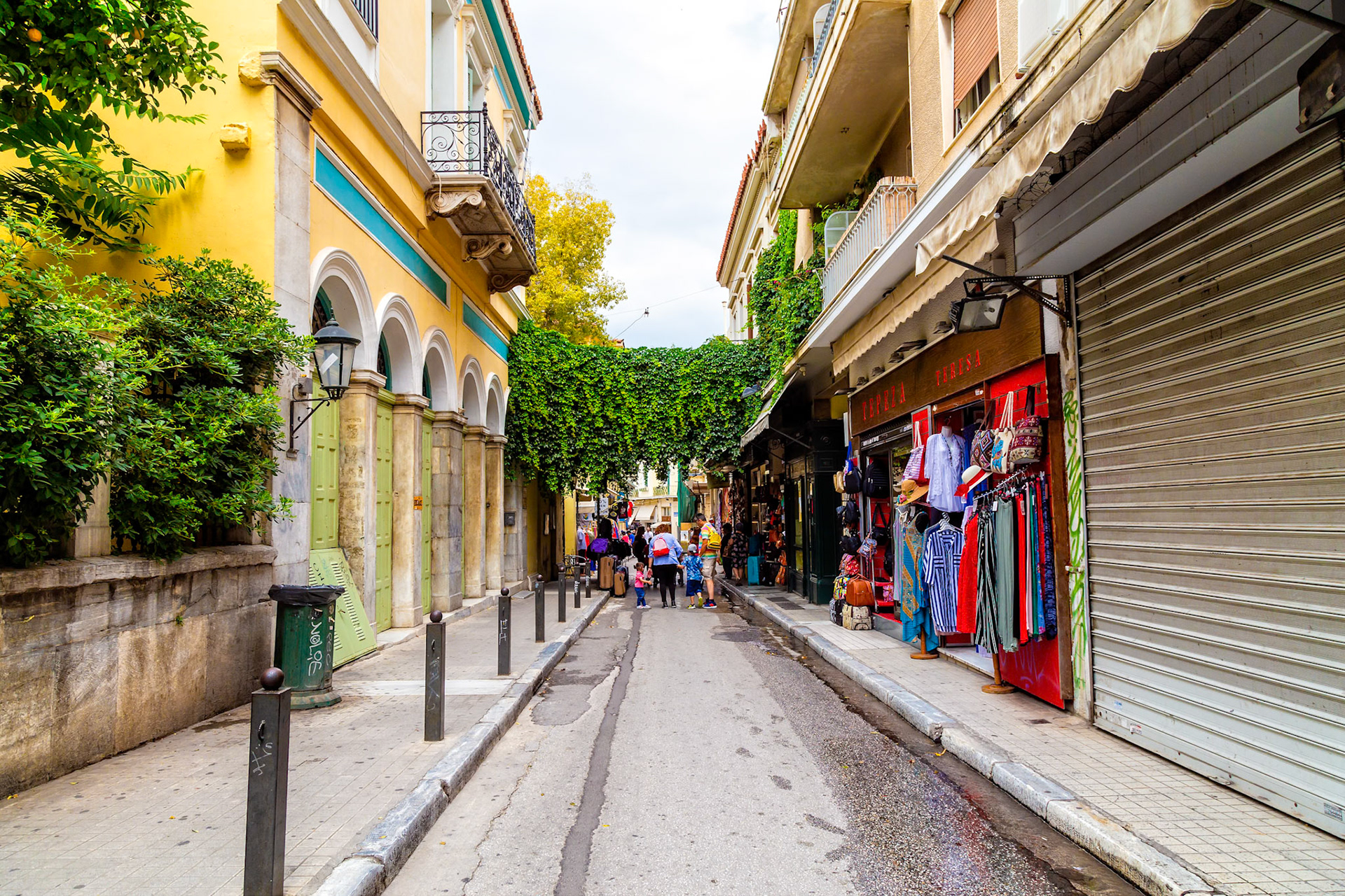 Athens, Greece - May 23rd 2018: Tourists stroll down a narrow street lined with shops, some closed, seeking souvenirs and experiencing the local culture.