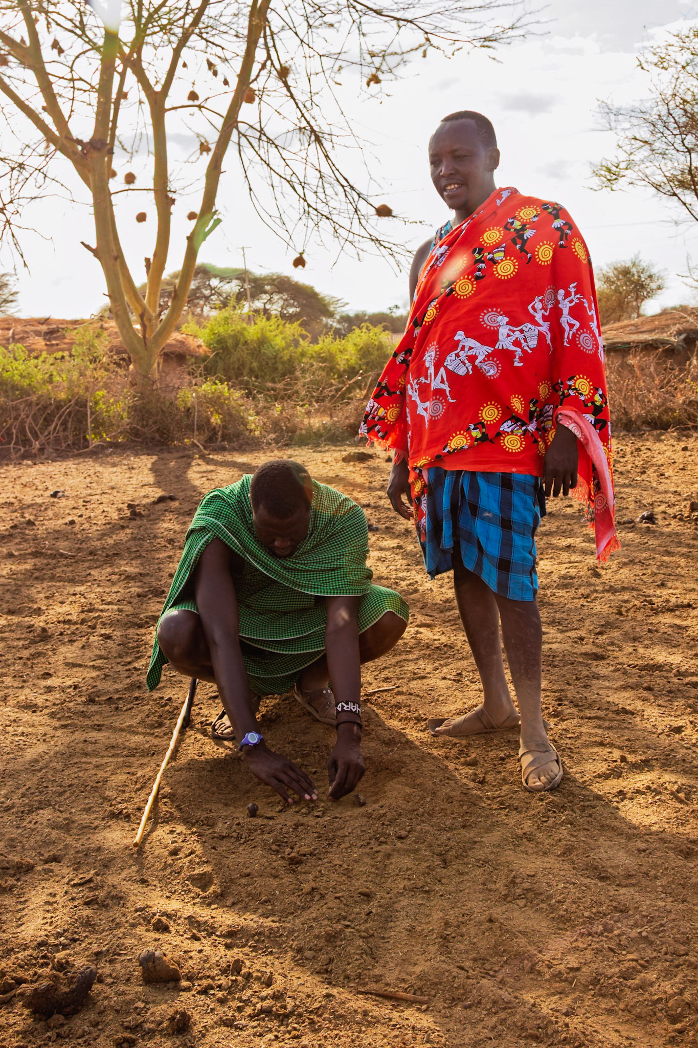 Two Maasai men in Kenya play a demonstrate firemaking skills in their village, wearing colorful shukas.