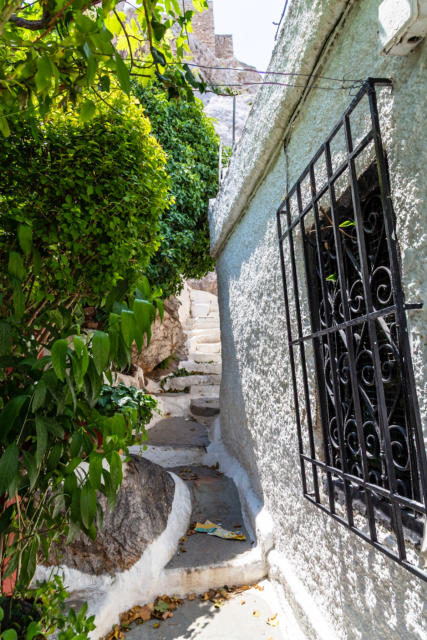 Athens, Greece - May 23rd 2018: A narrow, white-washed staircase ascends a hill, flanked by lush greenery and a building with a barred window, inviting exploration.