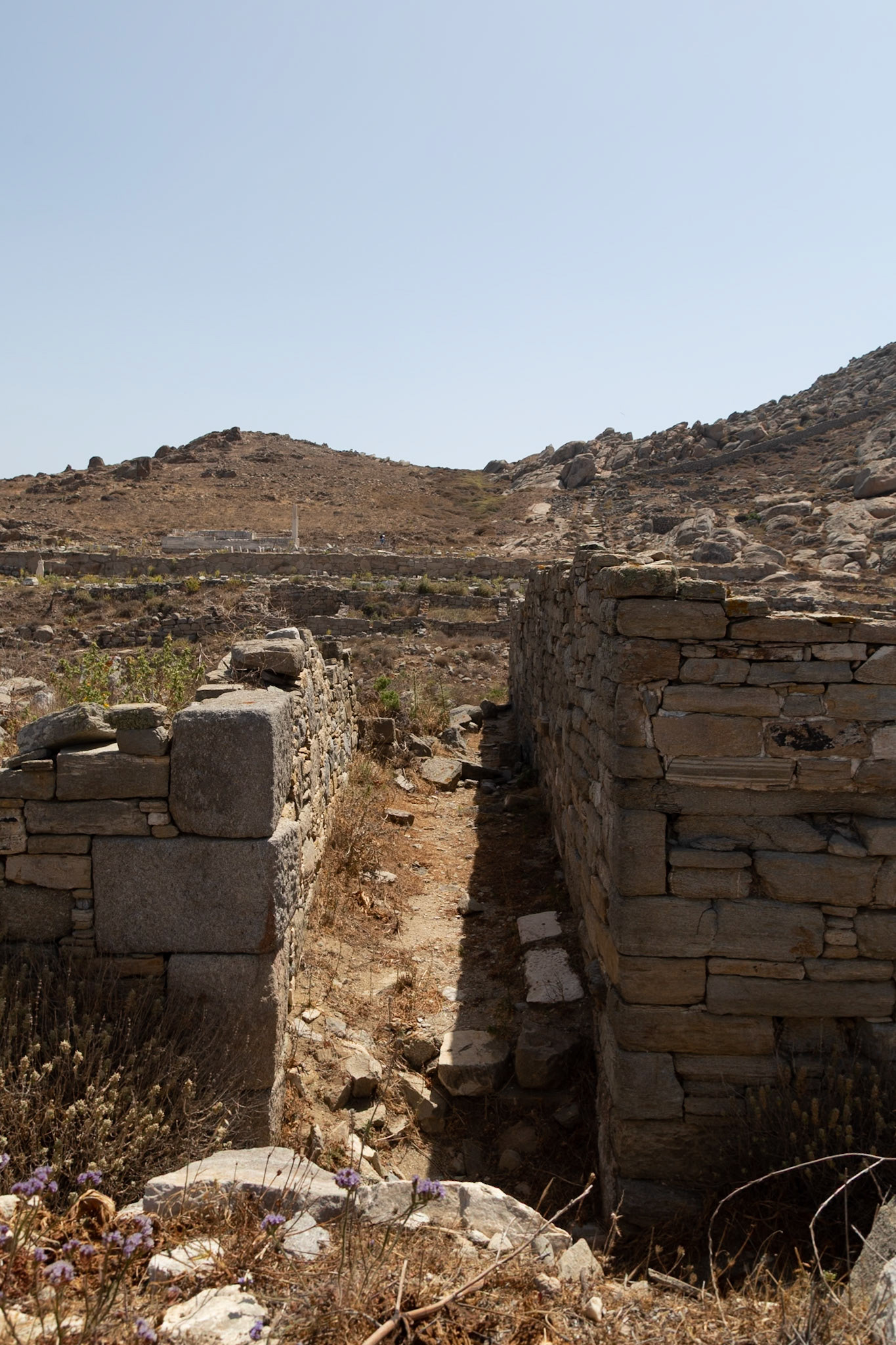 Delos, Greece - May 22nd 2018: A narrow path between ancient stone walls leads towards the ruins of Delos, a UNESCO World Heritage site, showcasing its rich history.