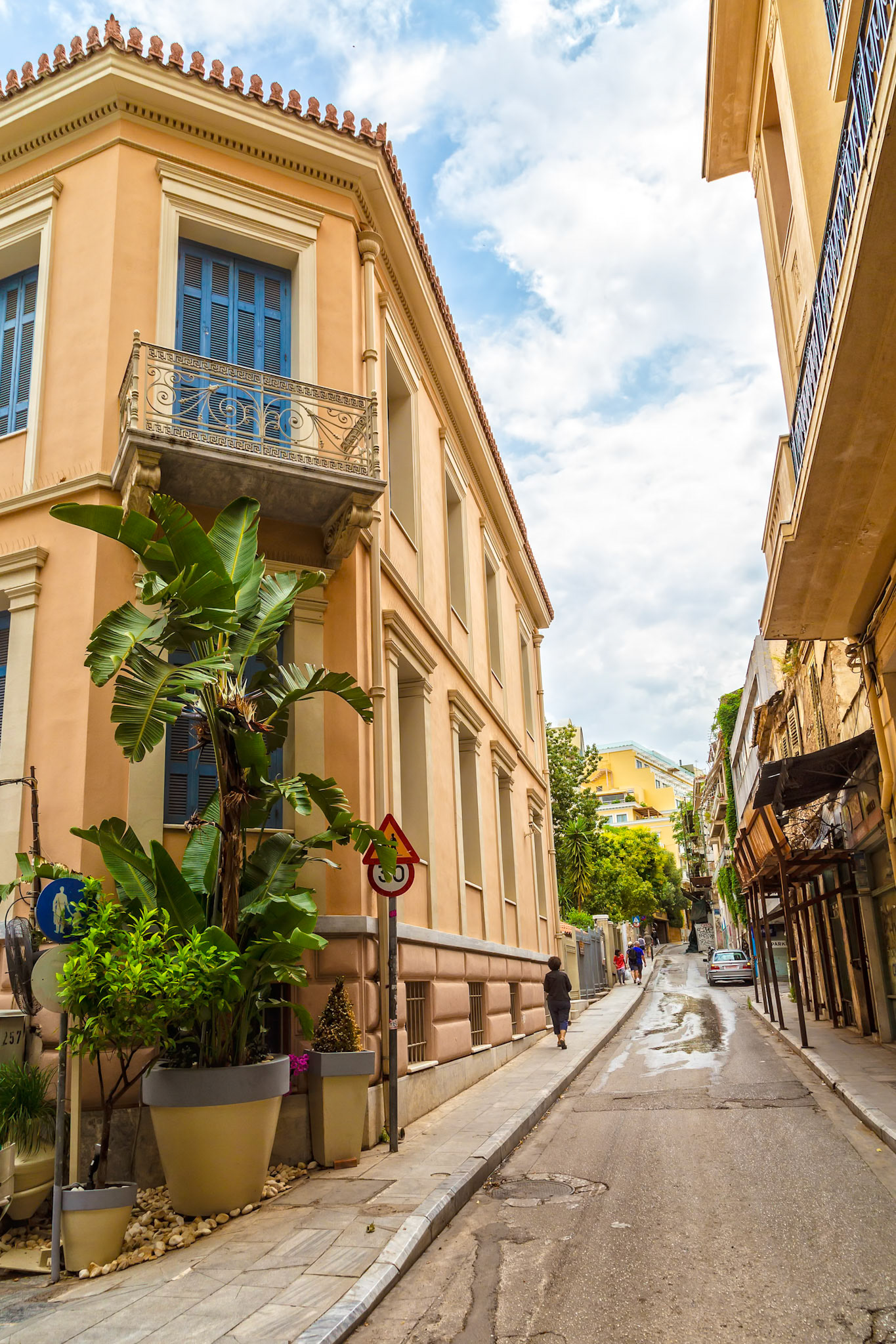 Athens, Greece - May 23rd 2018: Pedestrians walk along a narrow street lined with buildings and greenery, showcasing the urban landscape of Athens.
