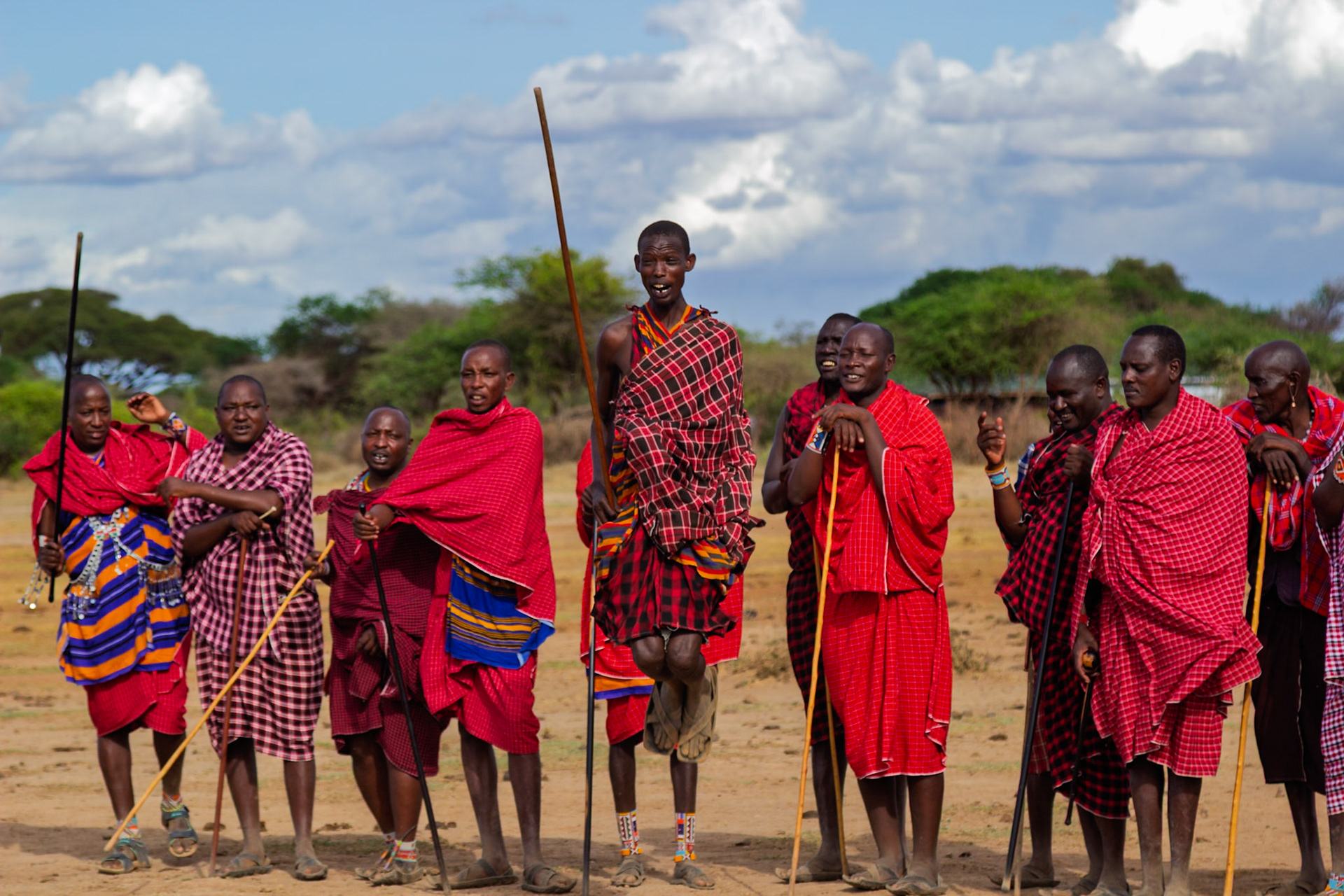 Maasai men in Kenya gather, one jumping with a stick, showcasing their traditional attire and cultural practices in their village.