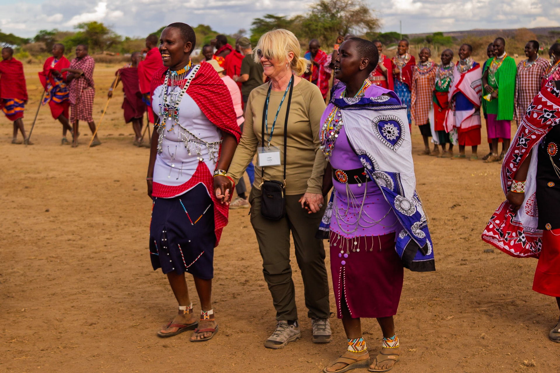 A tourist named Laura holds hands with Maasai women in traditional dress in a village in Kenya, experiencing their culture.