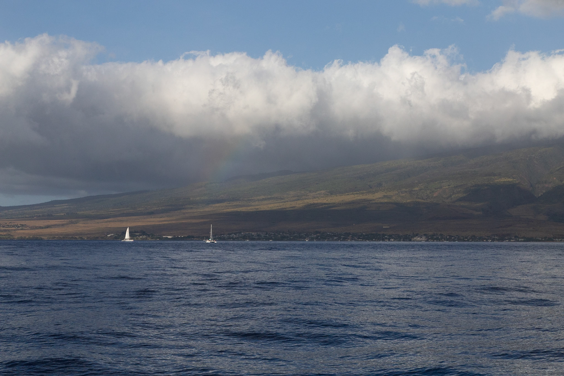Maui, Hawaii, USA - April 7th 2022: Sailboats are sailing on the ocean near the coast of Maui, with a rainbow appearing in the clouds above.