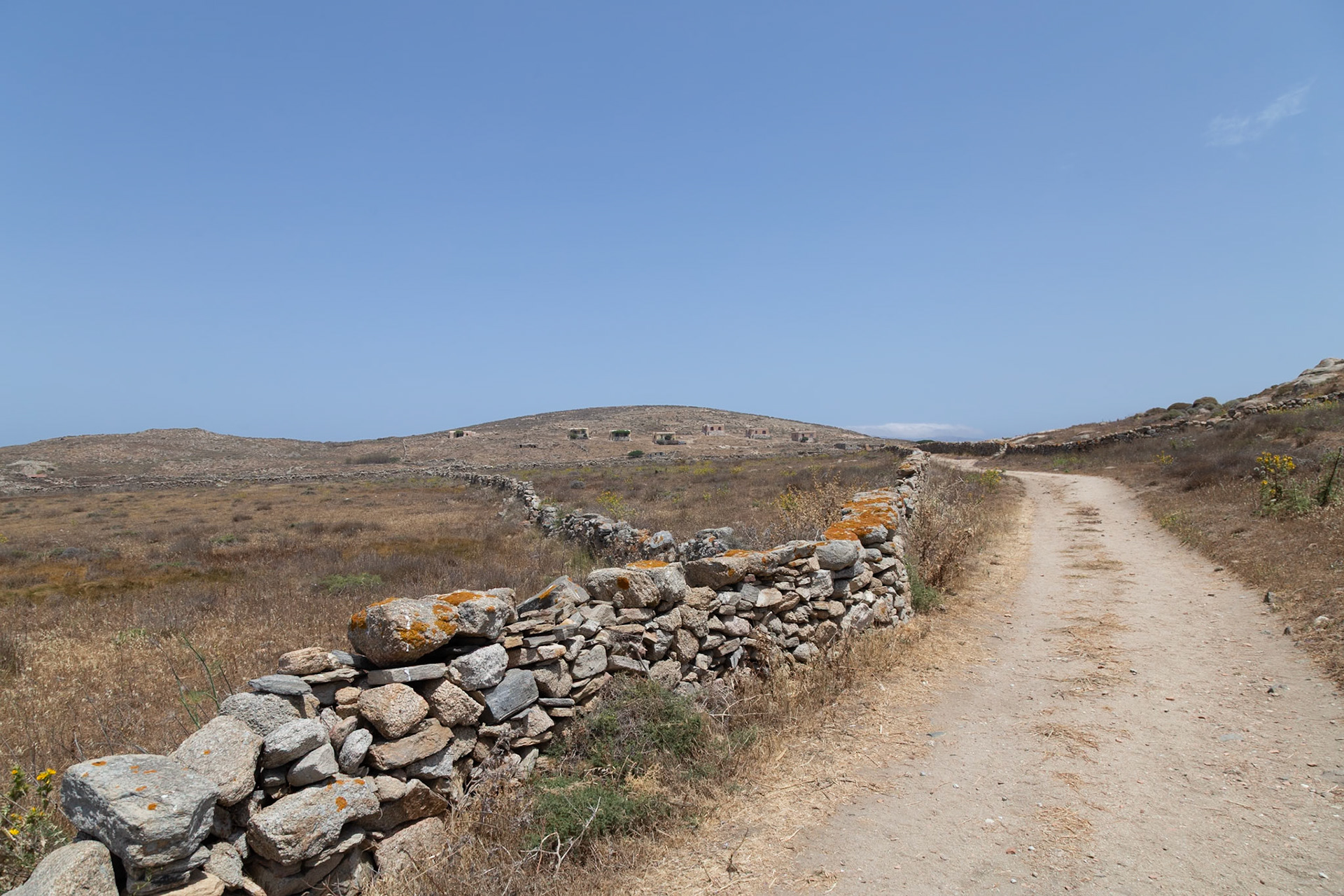 Delos, Greece - May 22nd 2018: A dirt path leads past a stone wall towards ruins on a hill. The scene captures the historical landscape of Delos.