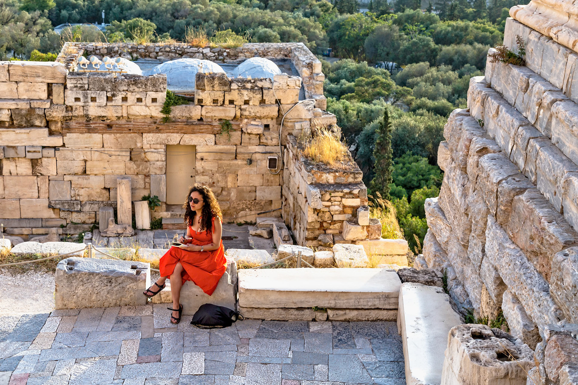 Acropolis, Athens, Greece - May 23rd 2018: A woman in a red dress sits on ancient stones, possibly sketching or writing, amidst the ruins of the Acropolis.