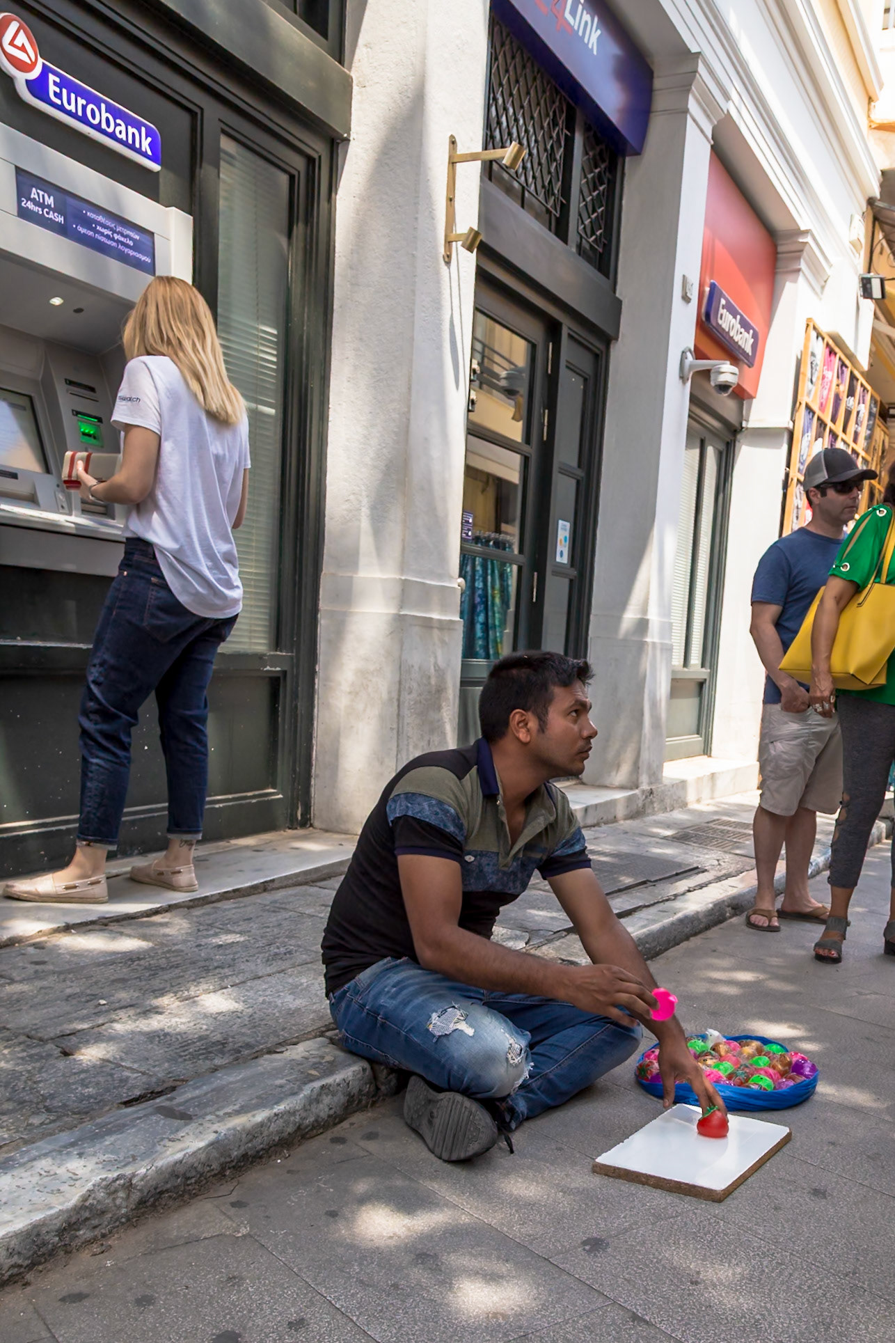 Athens, Greece - May 23rd 2018: A street vendor sits on the curb with toys, while a woman uses an ATM, and tourists walk by, capturing a slice of daily life.