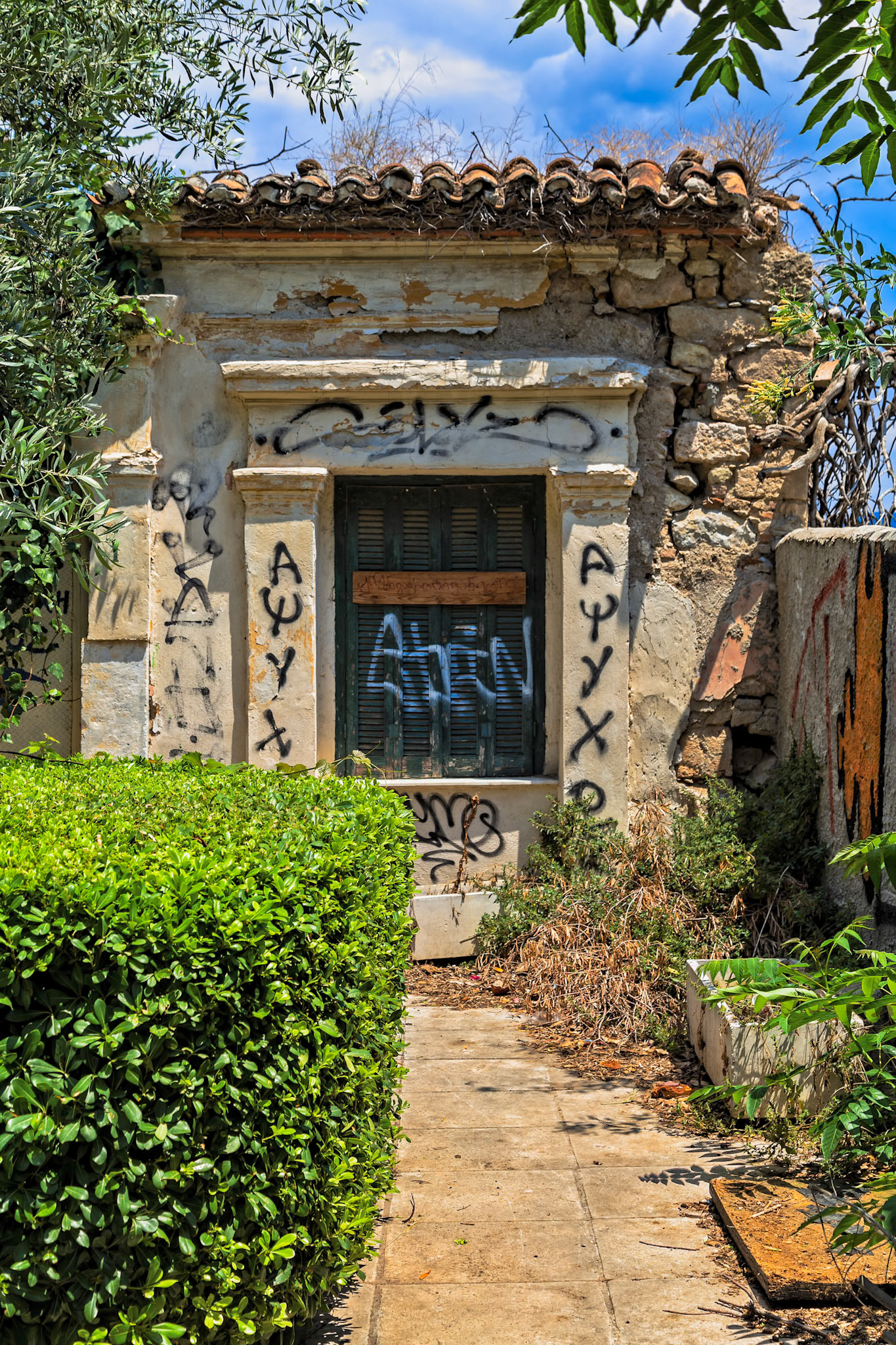 Athens, Greece - May 23rd 2018: An abandoned building with graffiti on the walls and boarded up windows sits overgrown with foliage.