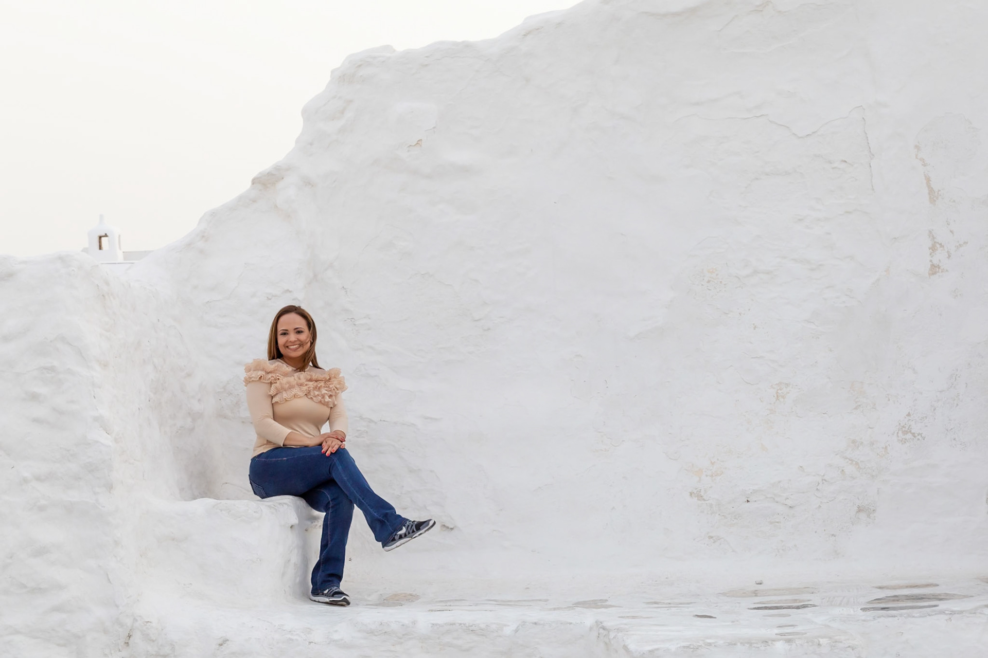 Mykonos, Greece - May 23rd 2018: A woman sits on a whitewashed wall, smiling for a photo. She is enjoying the sights of the Greek island.