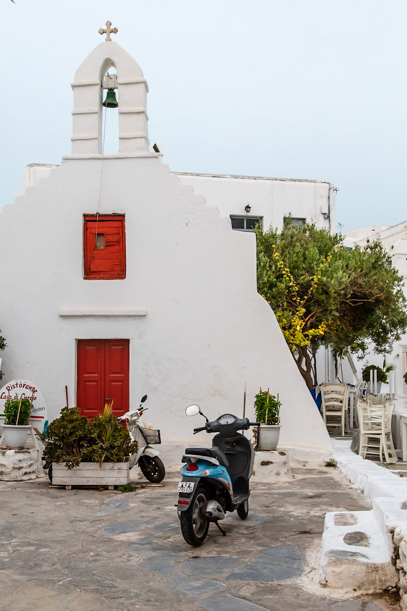 Mykonos, Greece - May 23rd 2018: A quaint chapel stands with scooters parked outside, near a restaurant, capturing the island's charm.