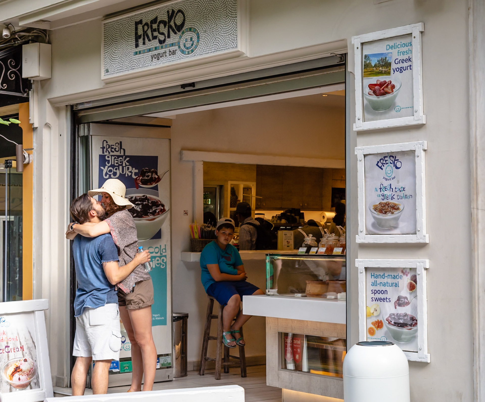 Athens, Greece - May 23rd 2018: A couple embraces outside Fresko Yogurt Bar, while a boy sits inside, waiting for a treat.