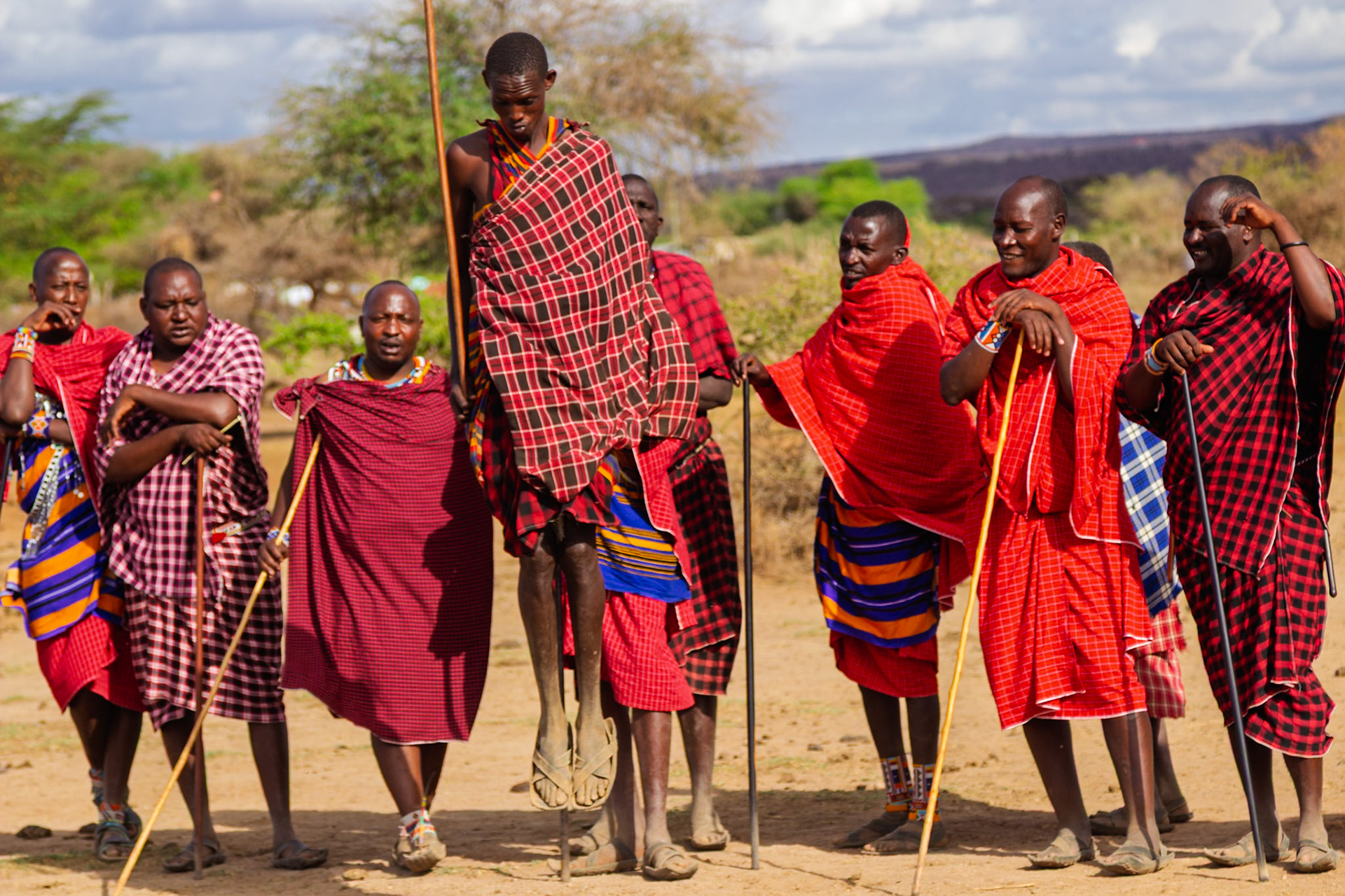 Maasai men in Kenya gather, one jumping high, showcasing their traditional attire and cultural practices in their village.