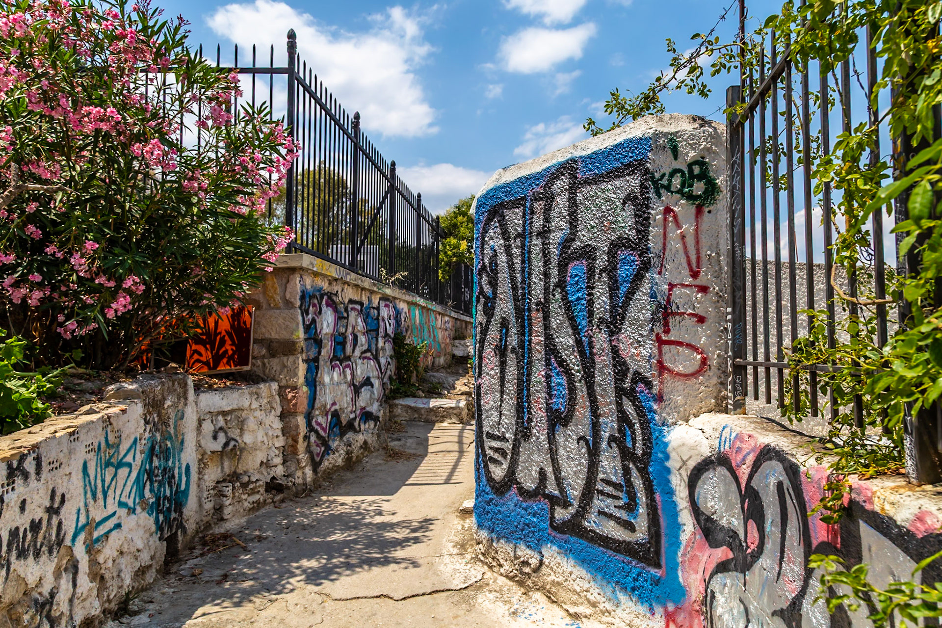 Athens, Greece - May 23rd 2018: A narrow alleyway is covered in graffiti, with a black fence and pink flowers adding color to the urban landscape.