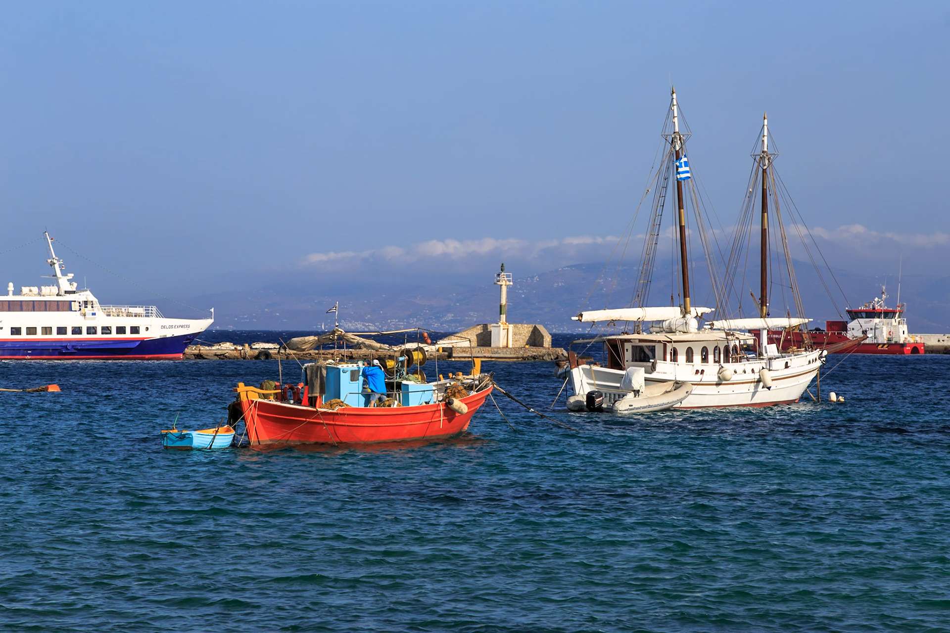 Mykonos, Greece - May 23rd 2018: Boats are moored in the harbor, including a red fishing boat and a white sailboat, awaiting their next voyage.