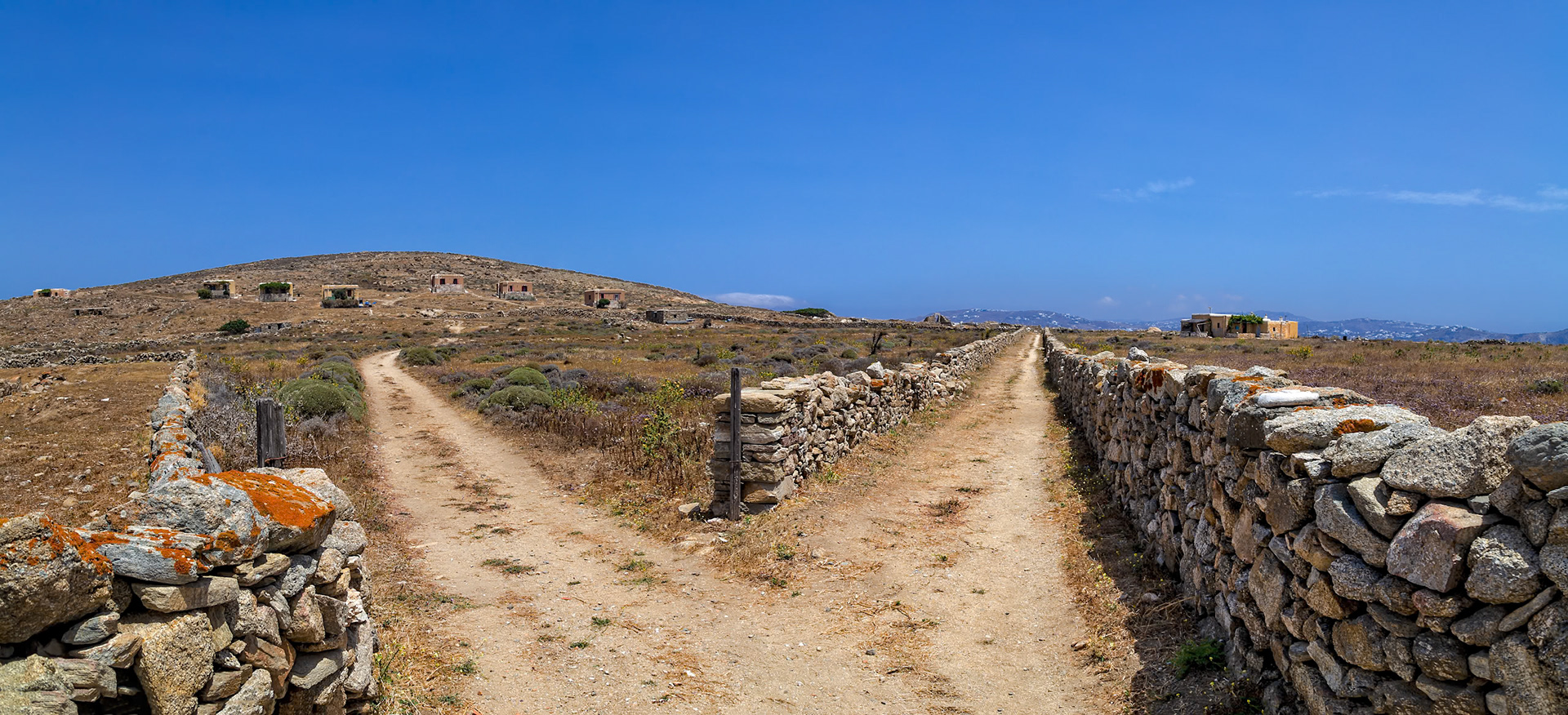 Delos, Greece - May 22nd 2018: A dirt path forks in Delos, Greece, with stone walls on either side. The path leads to ruins on a hill, showcasing the island's ancient history.