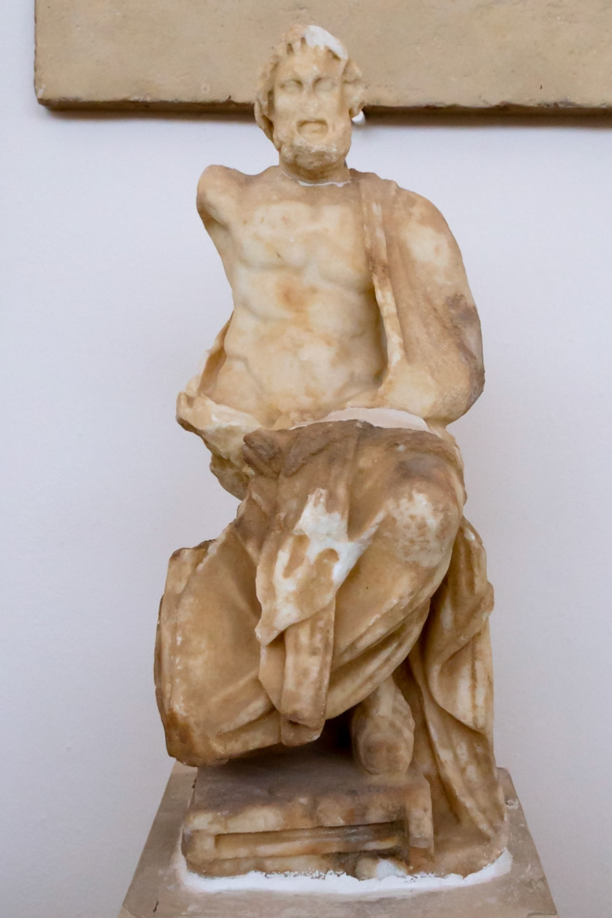 Delos, Greece - May 22nd 2018: A weathered marble statue of a male figure, possibly a god or hero, stands on display, showcasing ancient Greek artistry.