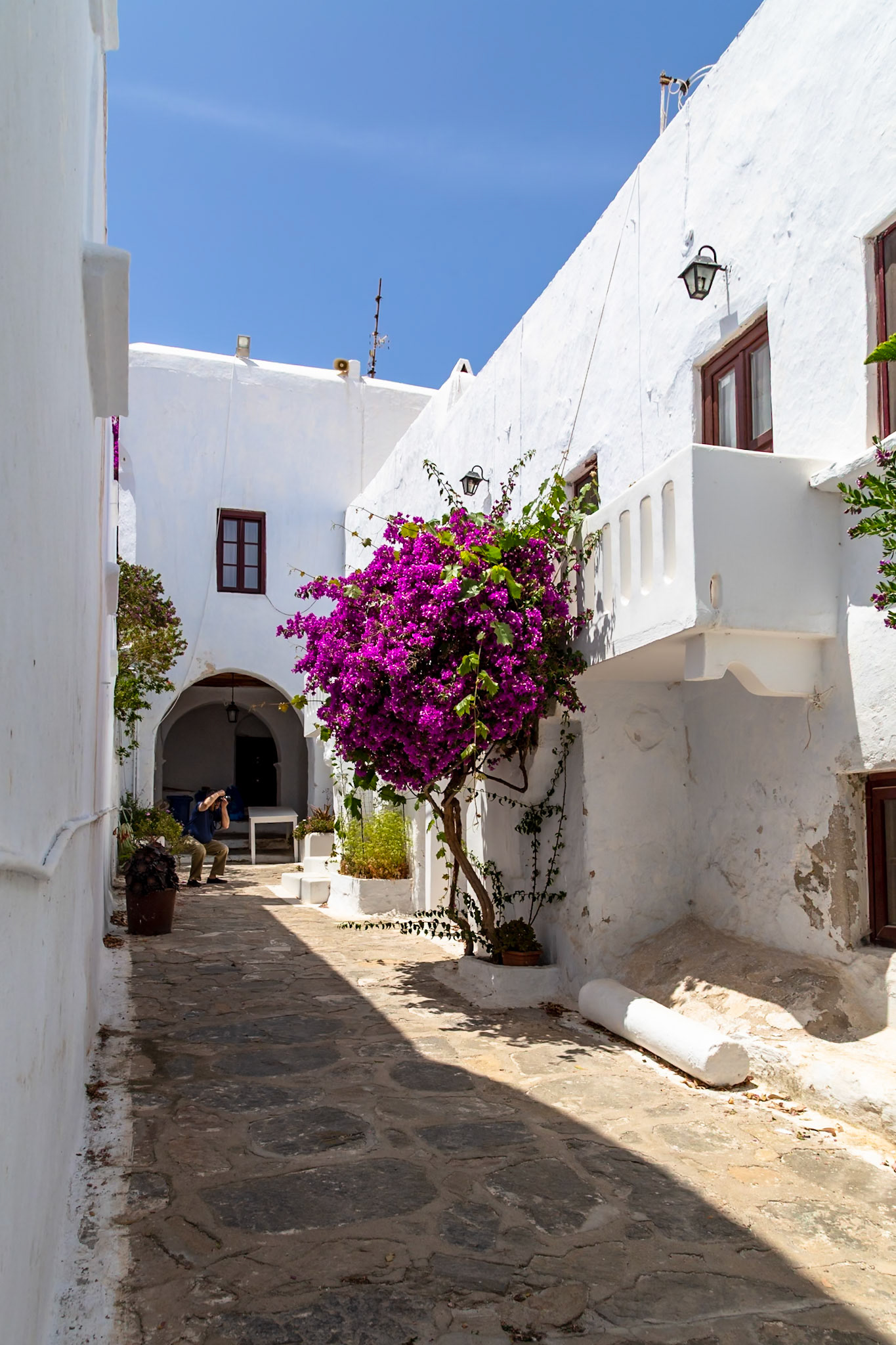 Mykonos, Greece - May 22nd 2018: A tourist takes photos in a narrow street lined with white buildings and vibrant bougainvillea.