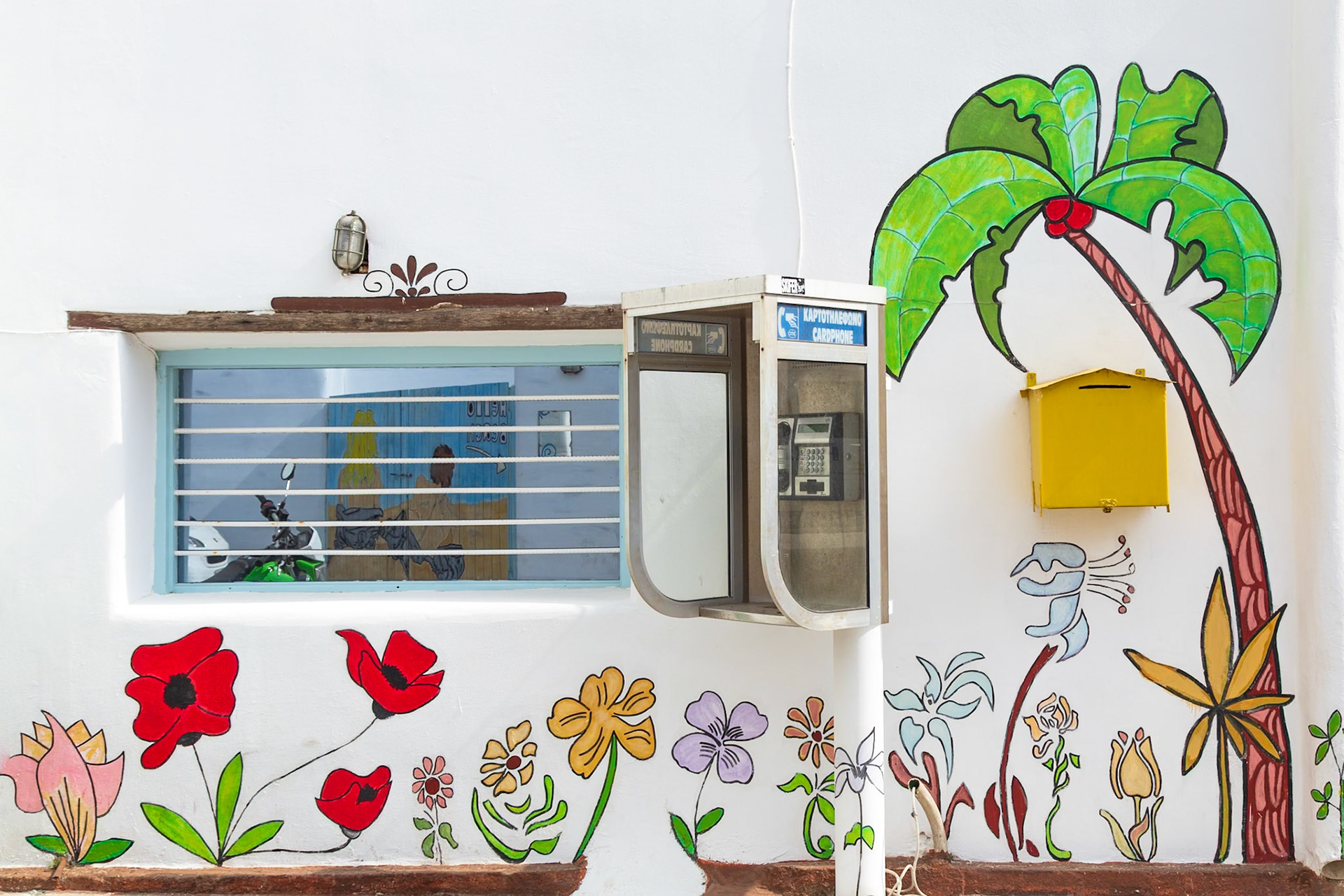 Paradise Beach, Mykonos, Greece - May 24th 2018: A colorful mural with flowers and a palm tree adorns a wall with a phone booth and window.