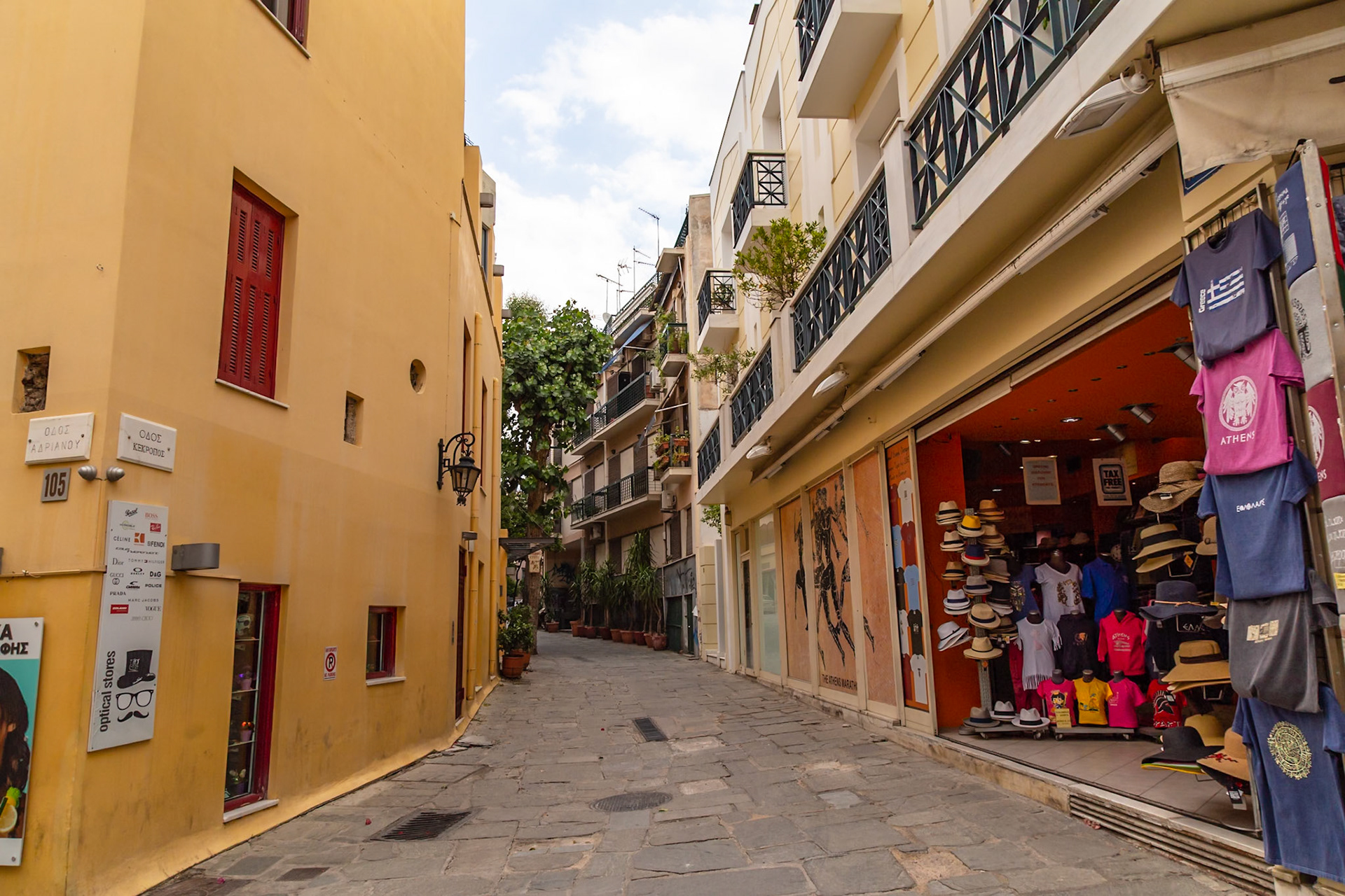 Athens, Greece - May 23rd 2018: A narrow street lined with shops and apartments. A souvenir shop is selling hats and t-shirts to tourists.