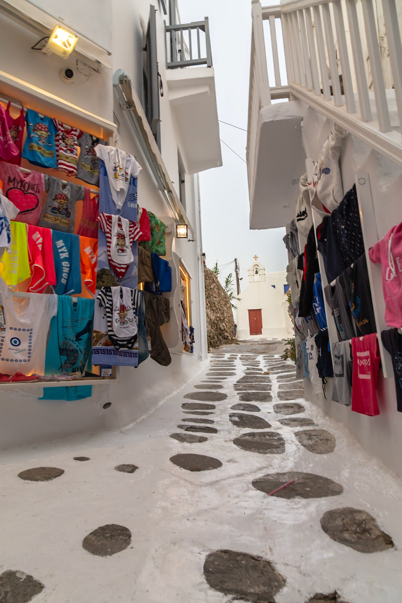 Mykonos, Greece - May 23rd 2018: A narrow street with shops selling clothes leads to a white church, showcasing the island's architecture.