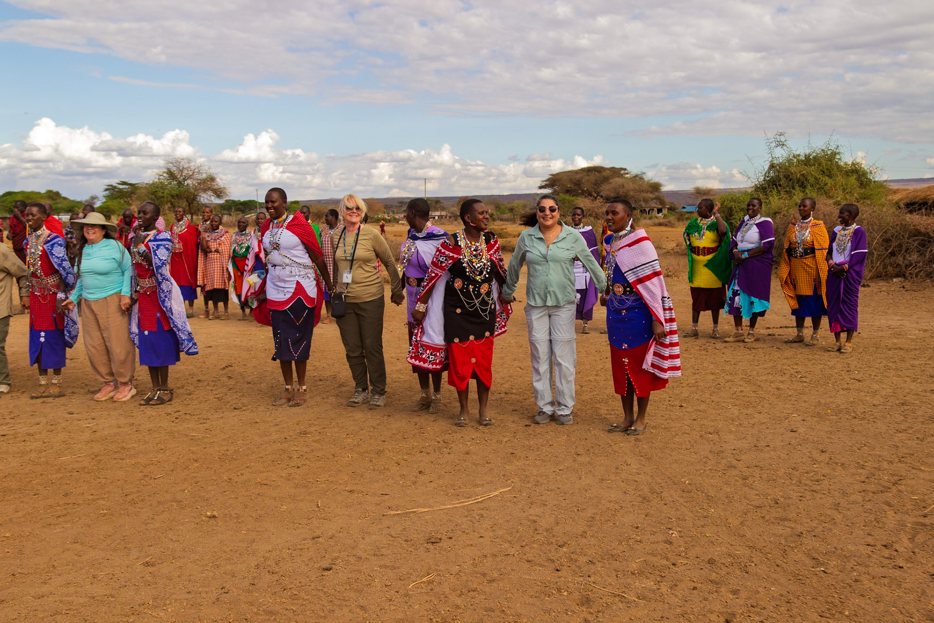 Tourists hold hands with Maasai women in traditional dress in a village in Kenya, experiencing local culture.