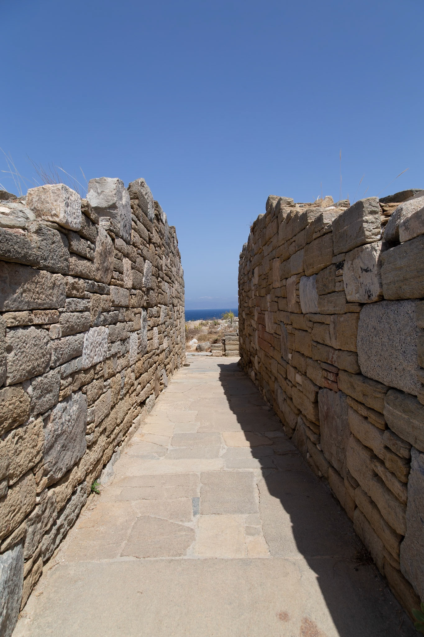 Delos, Greece - May 22nd 2018: A stone path between two walls leads to the sea. This is part of the ancient ruins of Delos, a UNESCO World Heritage site, attracting tourists.