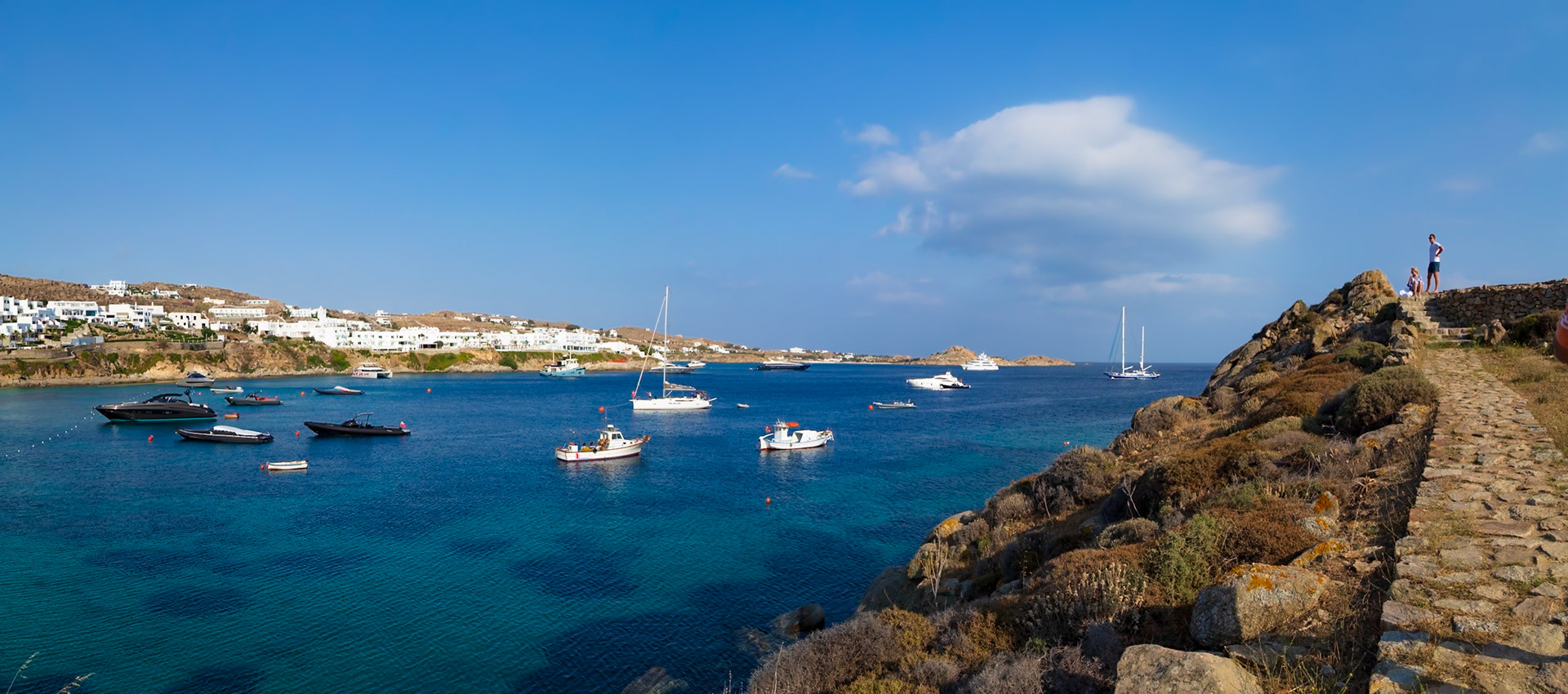 Psarou Beach, Mykonos, Greece - May 24th 2018: Boats dot the turquoise waters as tourists admire the view from a stone path on the coast.