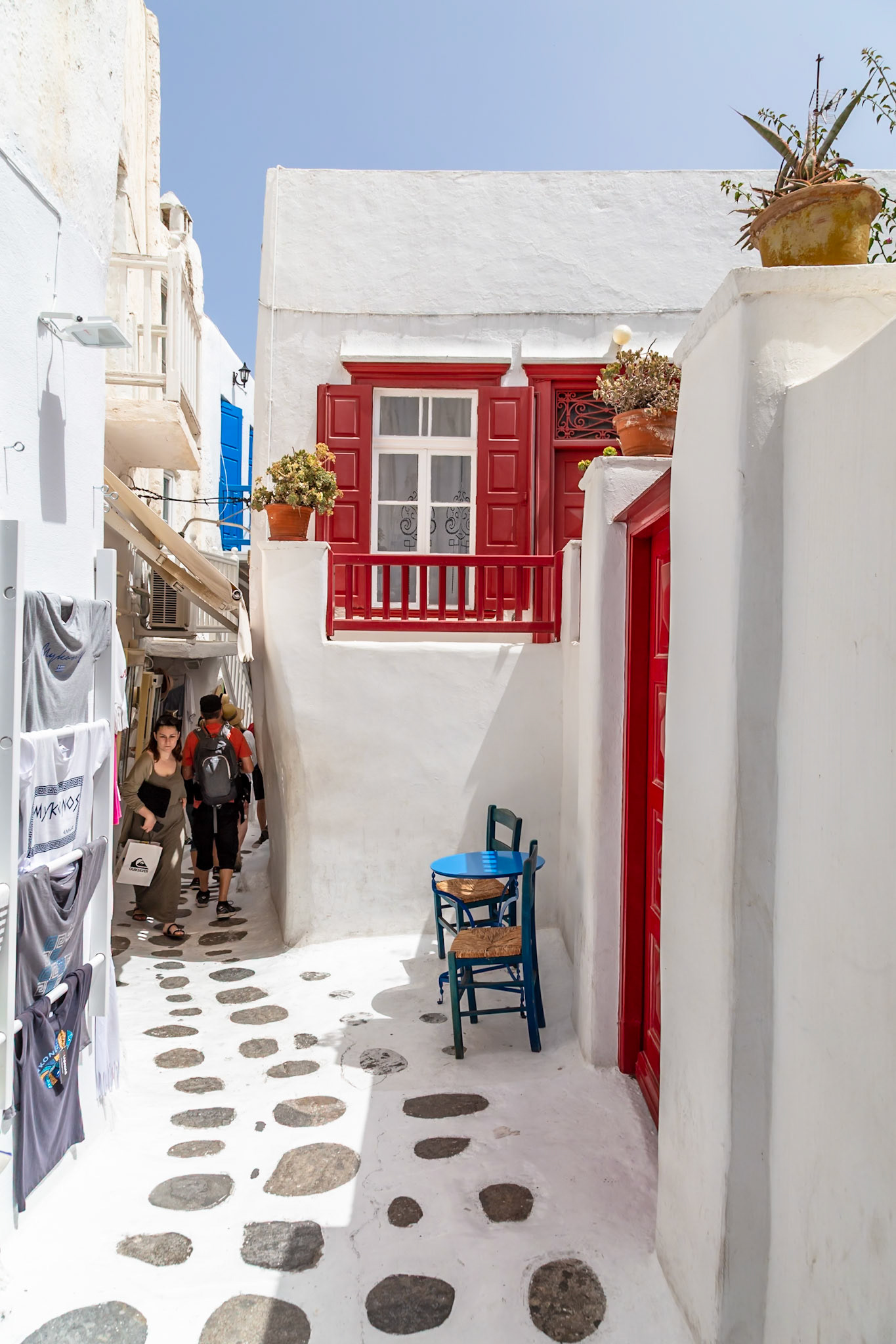 Mykonos, Greece - May 23rd 2018: Tourists stroll through a narrow, cobblestone street lined with white buildings and shops selling souvenirs.