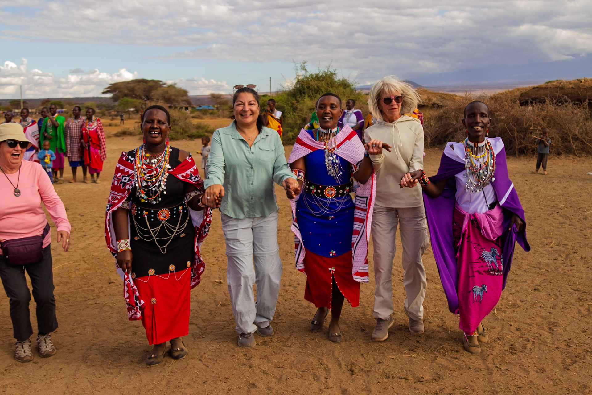 Tourists join Maasai women in Kenya, holding hands and walking together in a village. They are celebrating cultural exchange and friendship.