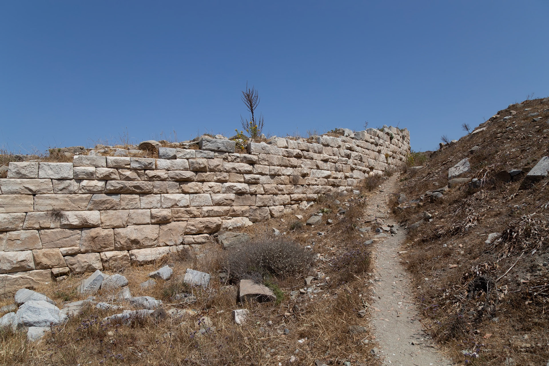 Delos, Greece - May 22nd 2018: An ancient stone wall stands beside a dirt path, showcasing the island's rich history and archaeological significance.