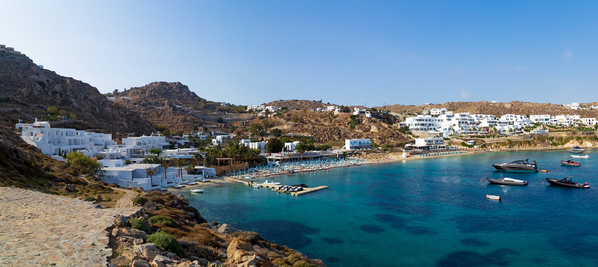 Psarou Beach, Mykonos, Greece - May 24th 2018: A scenic view of the beach with sunbeds, white buildings, and yachts in the turquoise water.