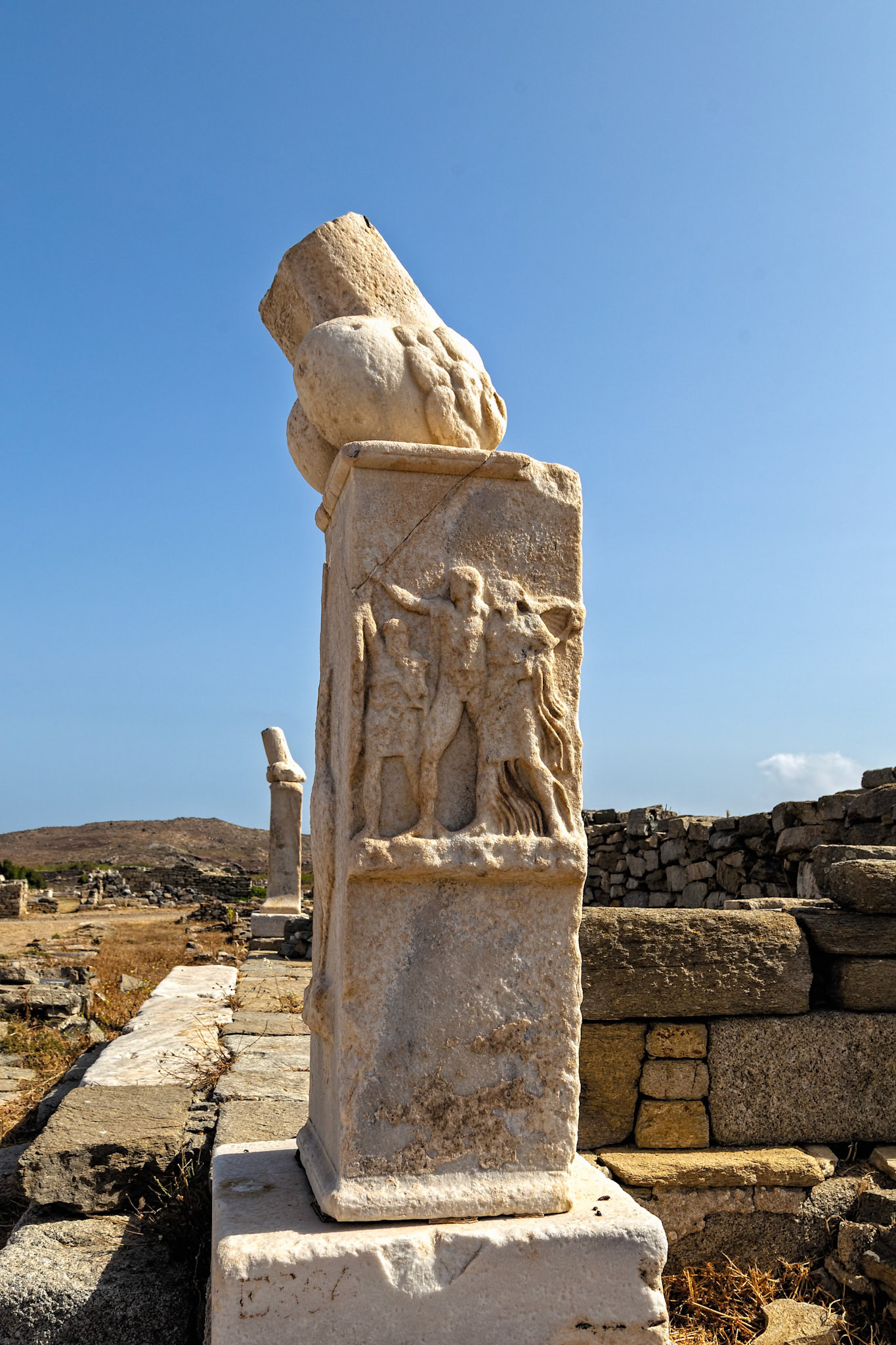 Delos, Greece - May 22nd 2018: A marble pillar with carved figures stands amidst the ruins of Delos, showcasing ancient Greek artistry and history.