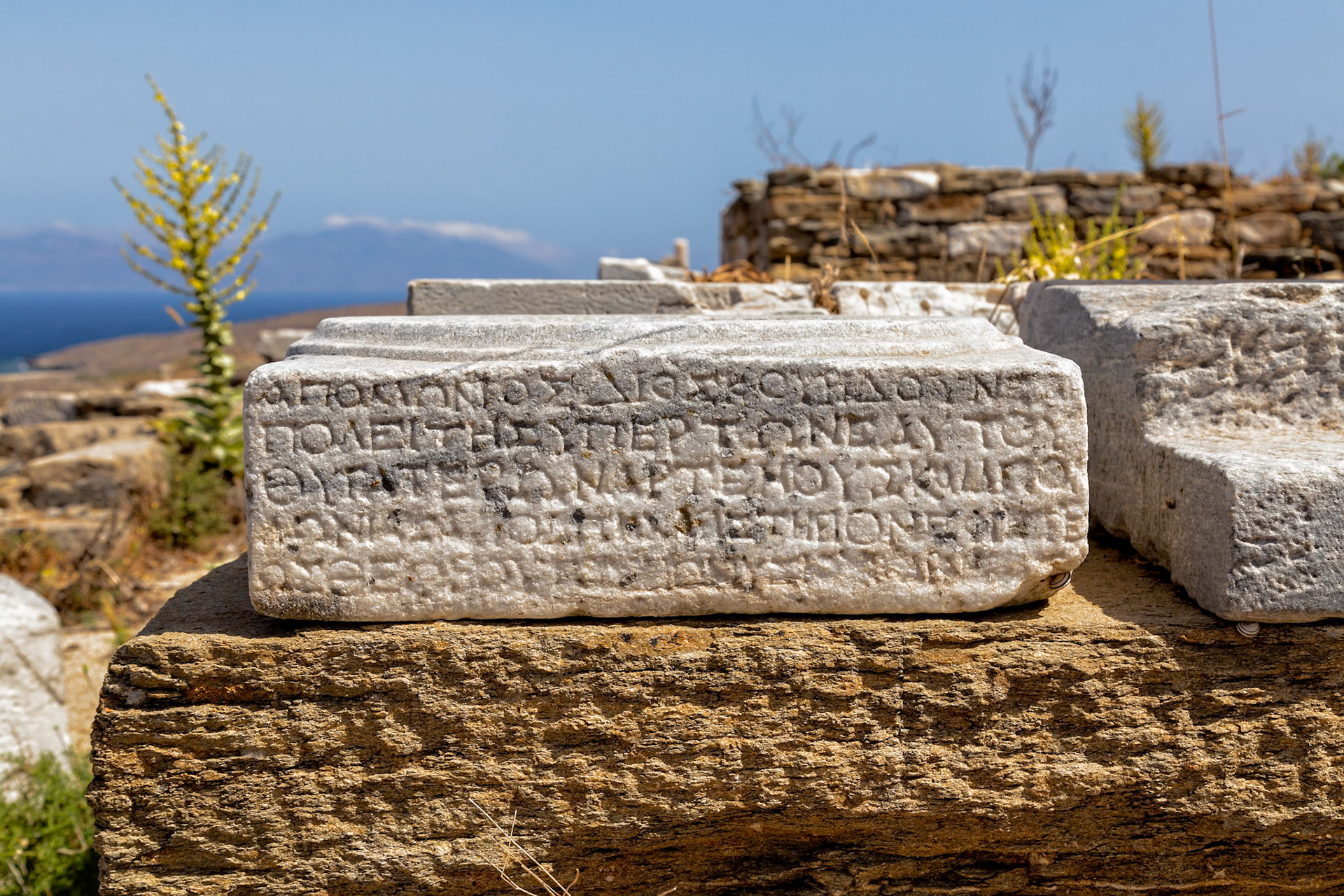 Delos, Greece - May 22nd 2018: An inscribed marble block rests atop a stone structure, showcasing ancient Greek text, likely part of a temple or public building.
