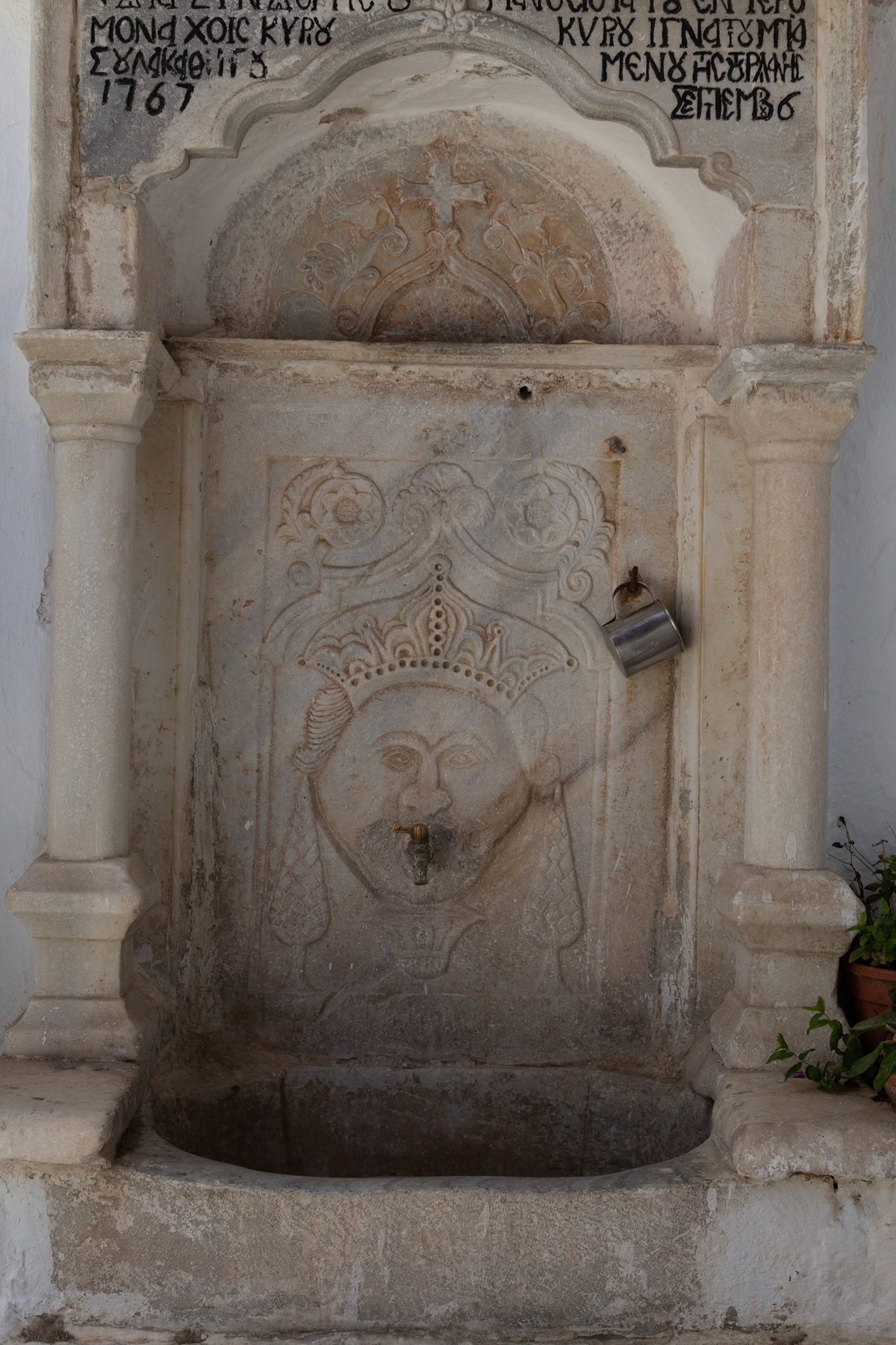 Mykonos, Greece - May 22nd 2018: An old stone water fountain with a carved face and a metal cup hanging on the side, likely for drinking.