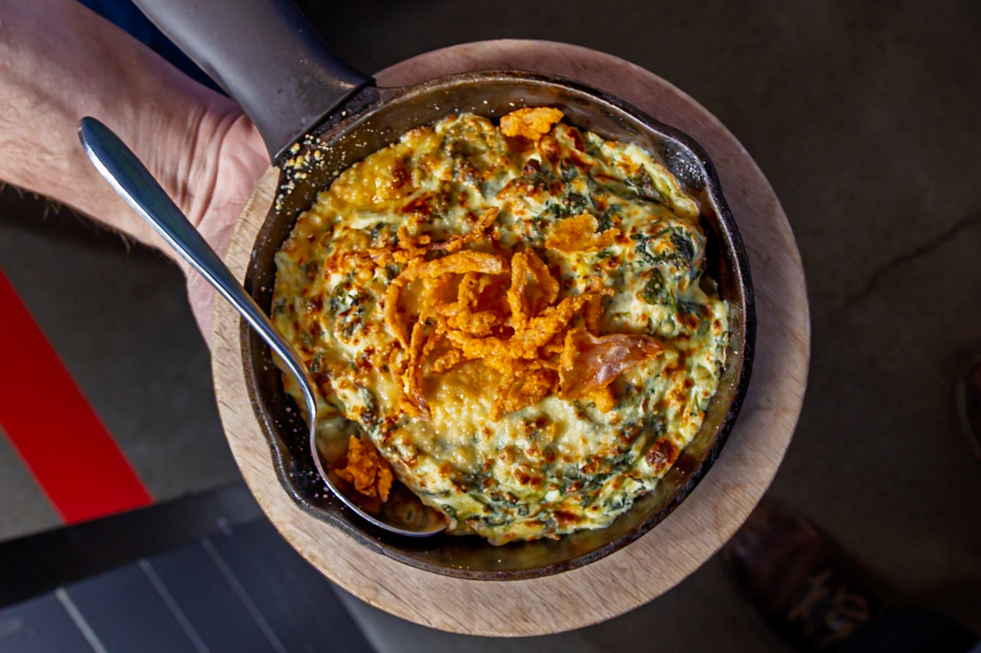 A person's hand holds a spoon over a cast iron skillet filled with a cheesy spinach dip topped with fried onions.