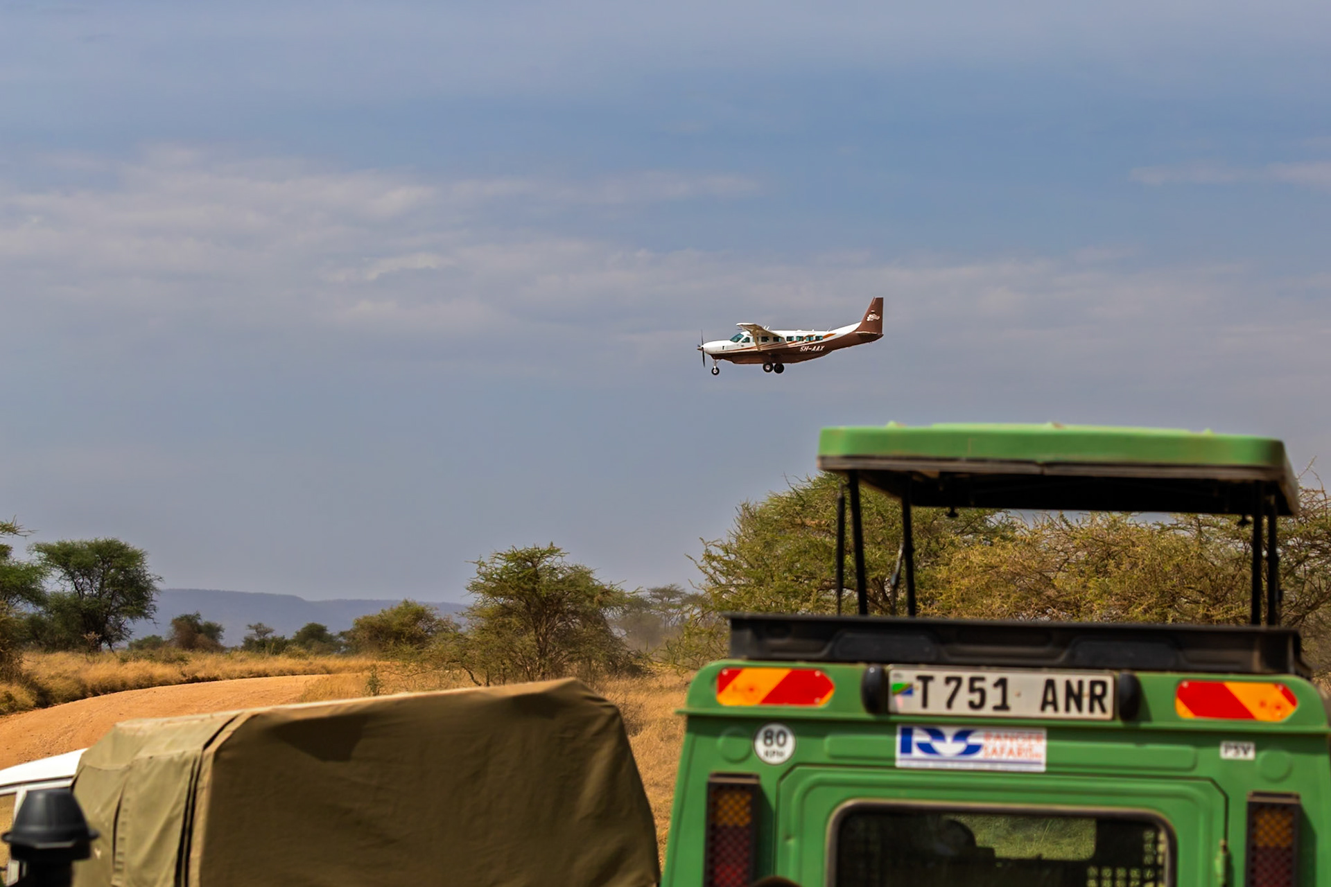 A plane flies over safari vehicles in Tanzania's Serengeti National Park, likely transporting tourists for wildlife viewing.