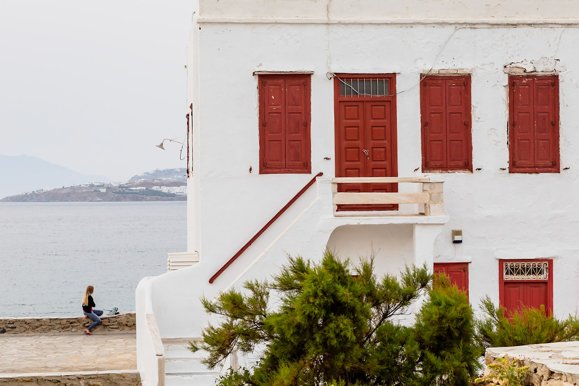 Mykonos, Greece - May 23rd 2018: A woman sits by the sea, enjoying the view of the town and the water, next to a white building with red doors.