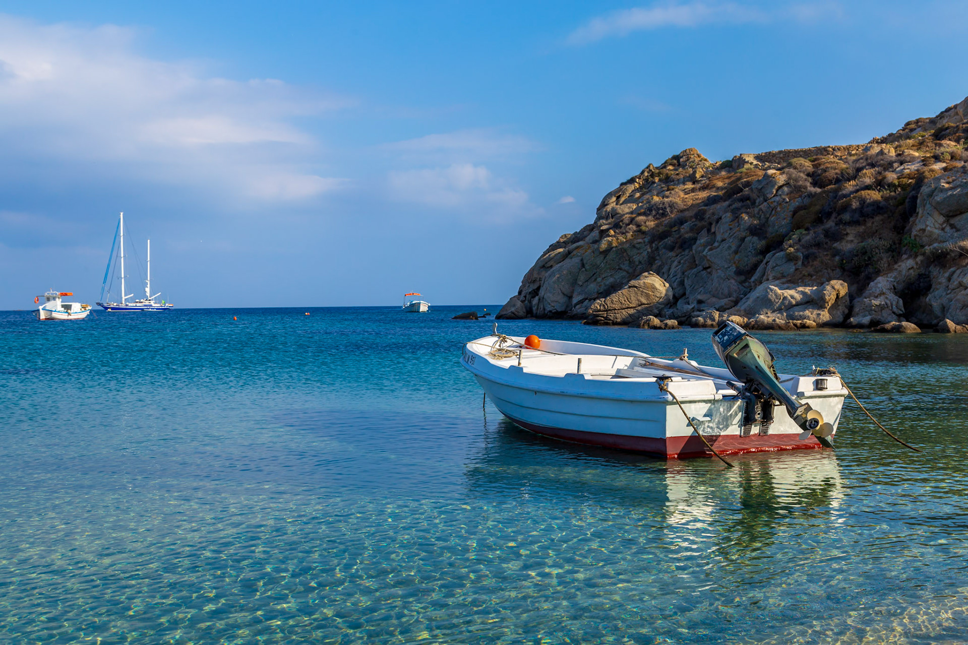Psarou Beach, Mykonos, Greece - May 24th 2018: A small motorboat rests in the clear, shallow water near the rocky shore, with other boats visible in the distance.