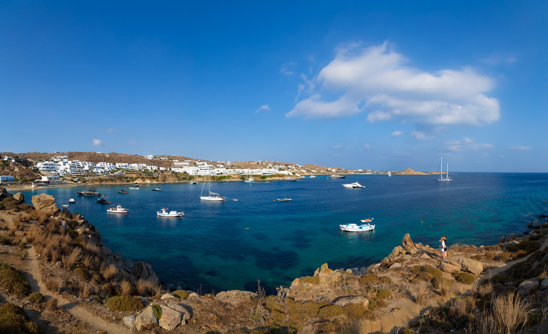 Psarou Beach, Mykonos, Greece - May 24th 2018: Boats dot the turquoise waters of Psarou Beach, while two people stand on the rocky cliffs, enjoying the view.