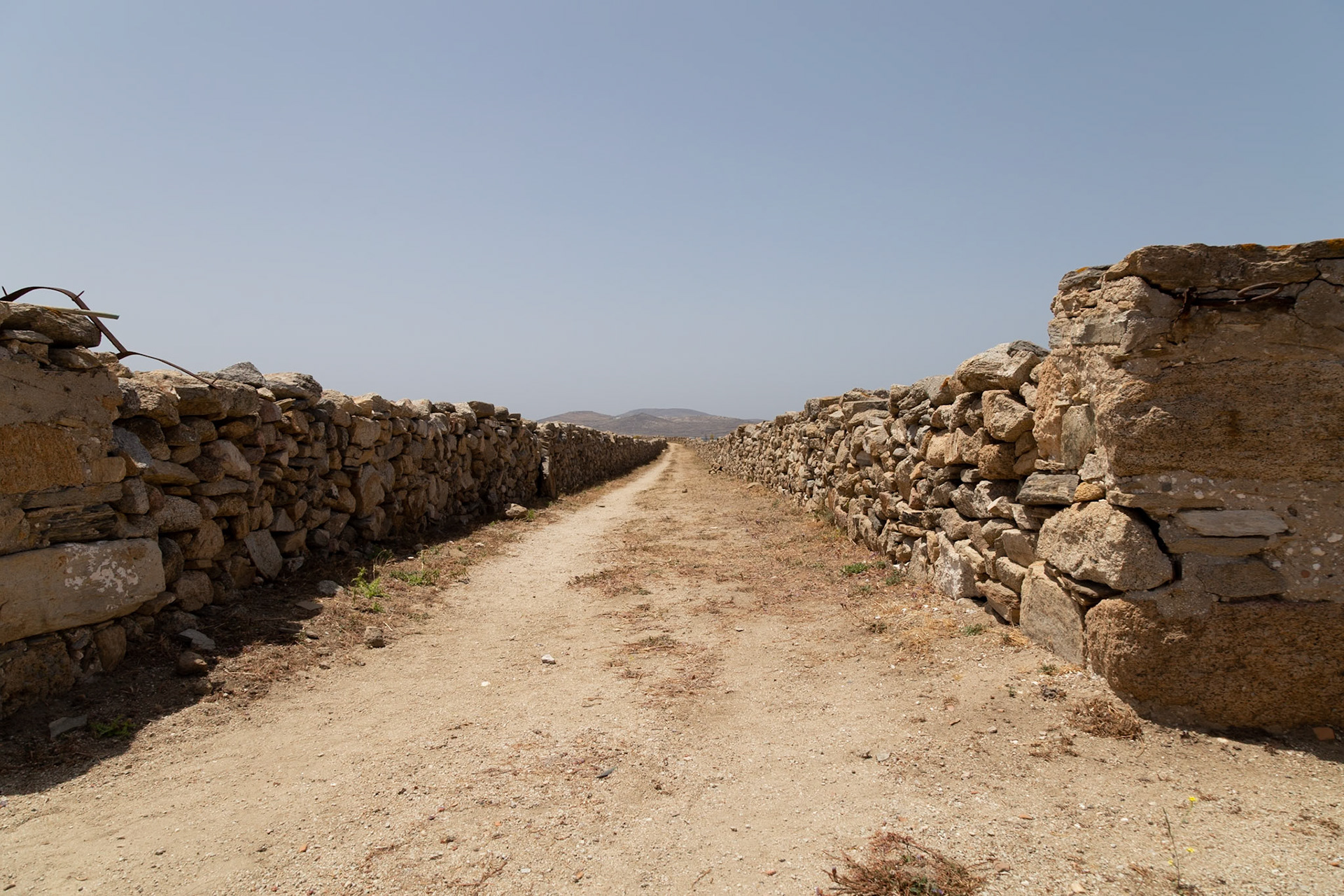 Delos, Greece - May 22nd 2018: A dirt path stretches between ancient stone walls, remnants of a once-thriving civilization, inviting exploration of Delos' rich history.