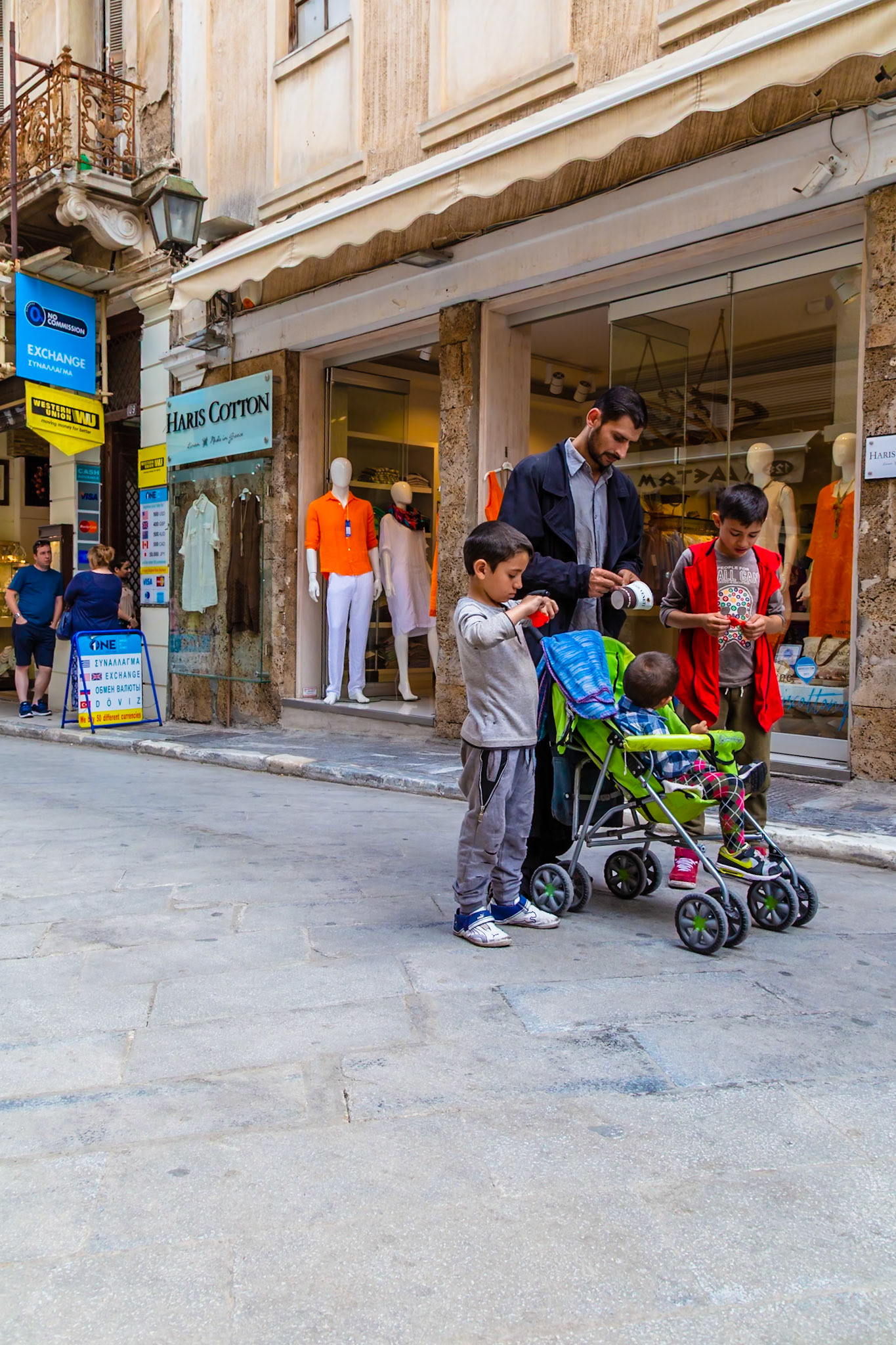 Athens, Greece - May 23rd 2018: A man is seen with two boys and a baby in a stroller on a city street, in front of the Haris Cotton store.