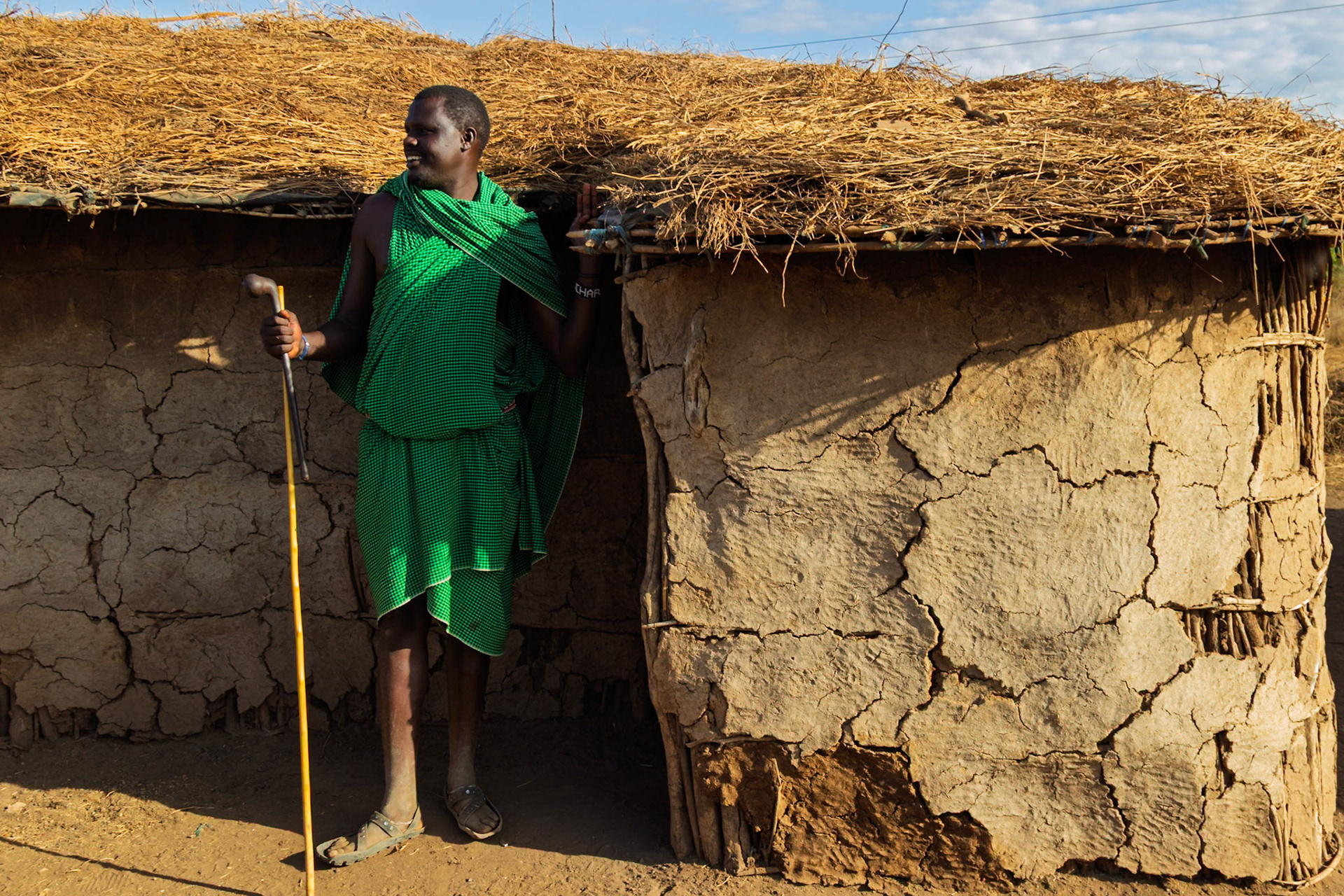 A Maasai man in Kenya stands outside a traditional mud hut, wearing a green shuka and holding a walking stick.