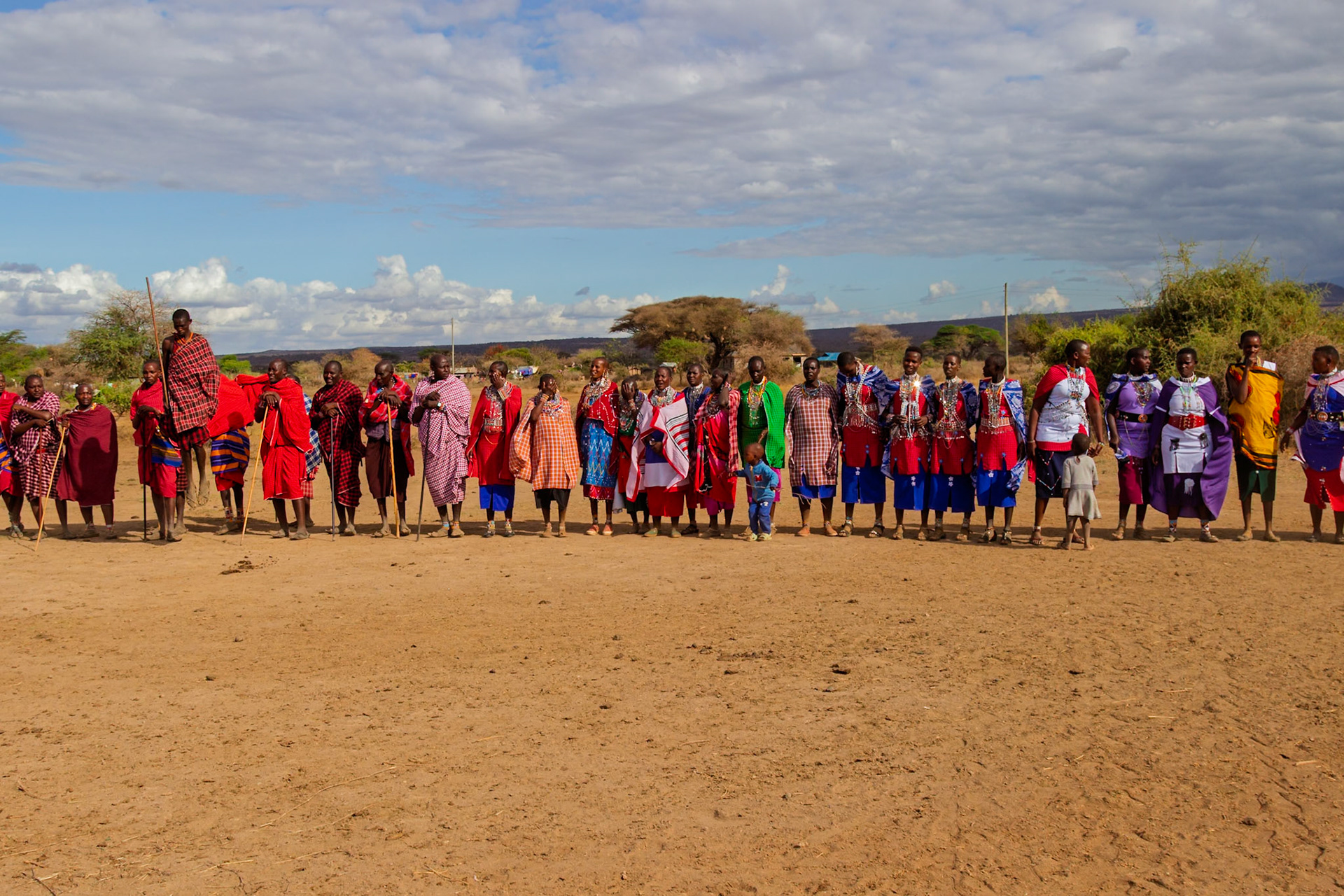 A group of Maasai people in Kenya are standing together in their village, wearing traditional clothing, for a group photo.