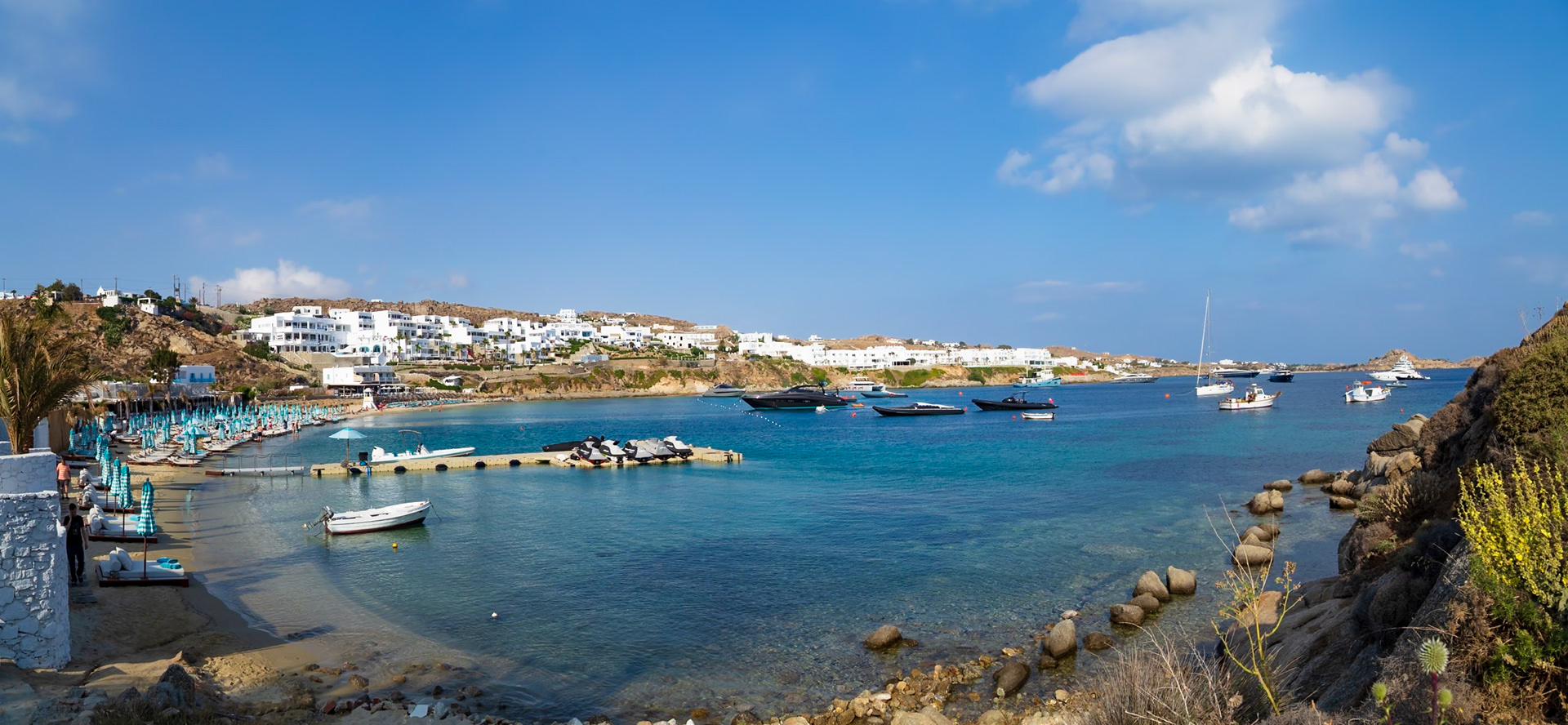 Psarou Beach, Mykonos, Greece - May 24th 2018: A scenic view of the beach with yachts and jet skis in the water, and beachgoers enjoying the sun.